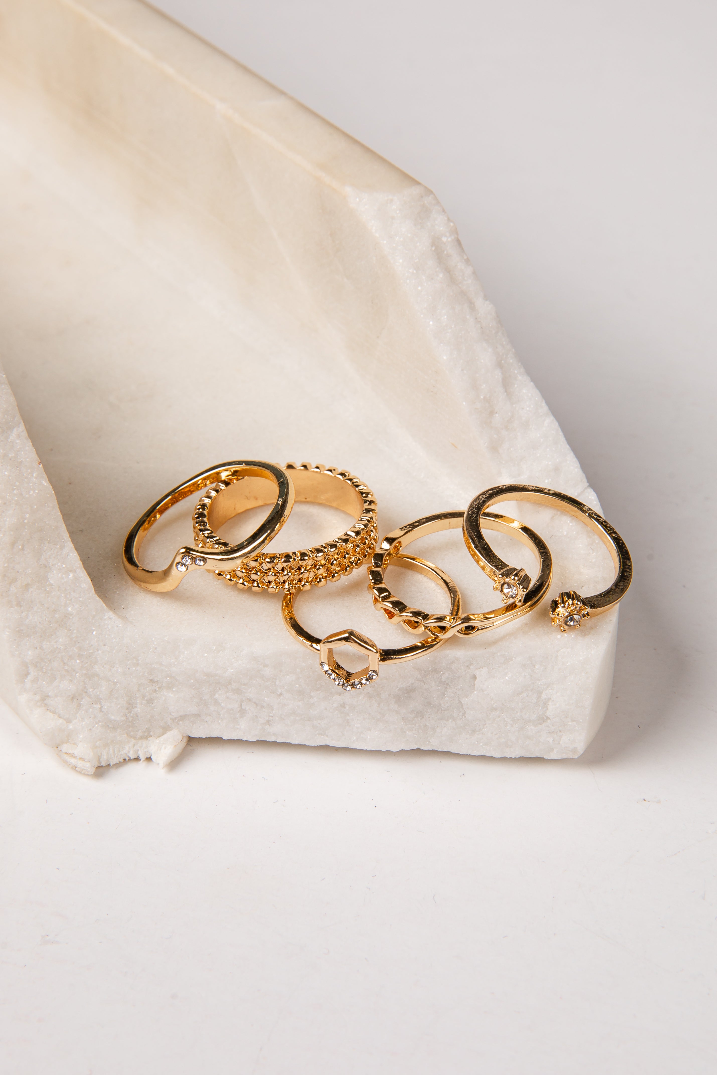 A set of five gold rings with various textures and designs, including a heart-shaped ring and a beaded ring, displayed on a white textured stone tray against a light gray background.