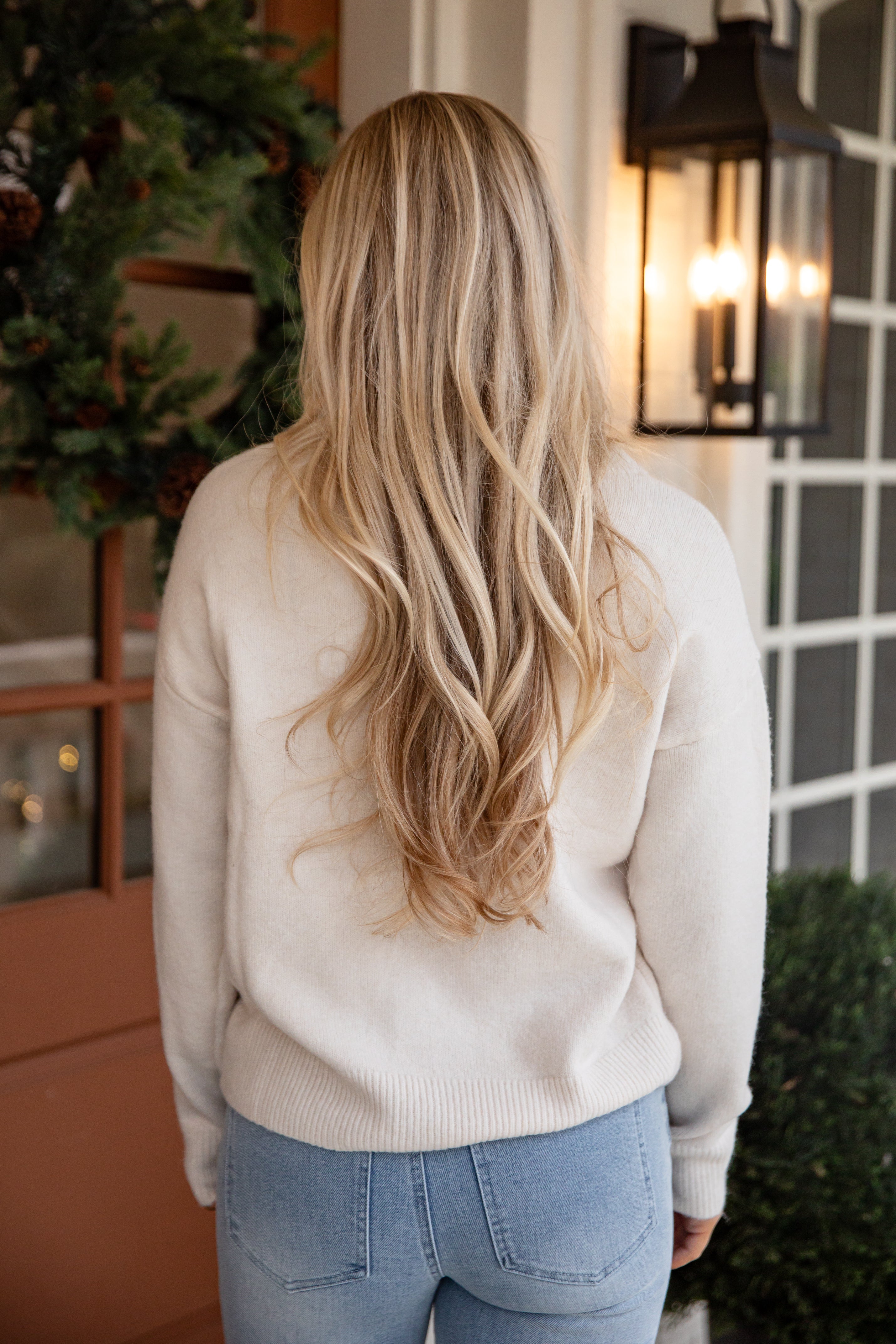 Person wearing a beige sweater and blue jeans with long blonde hair, standing in front of a house entrance.