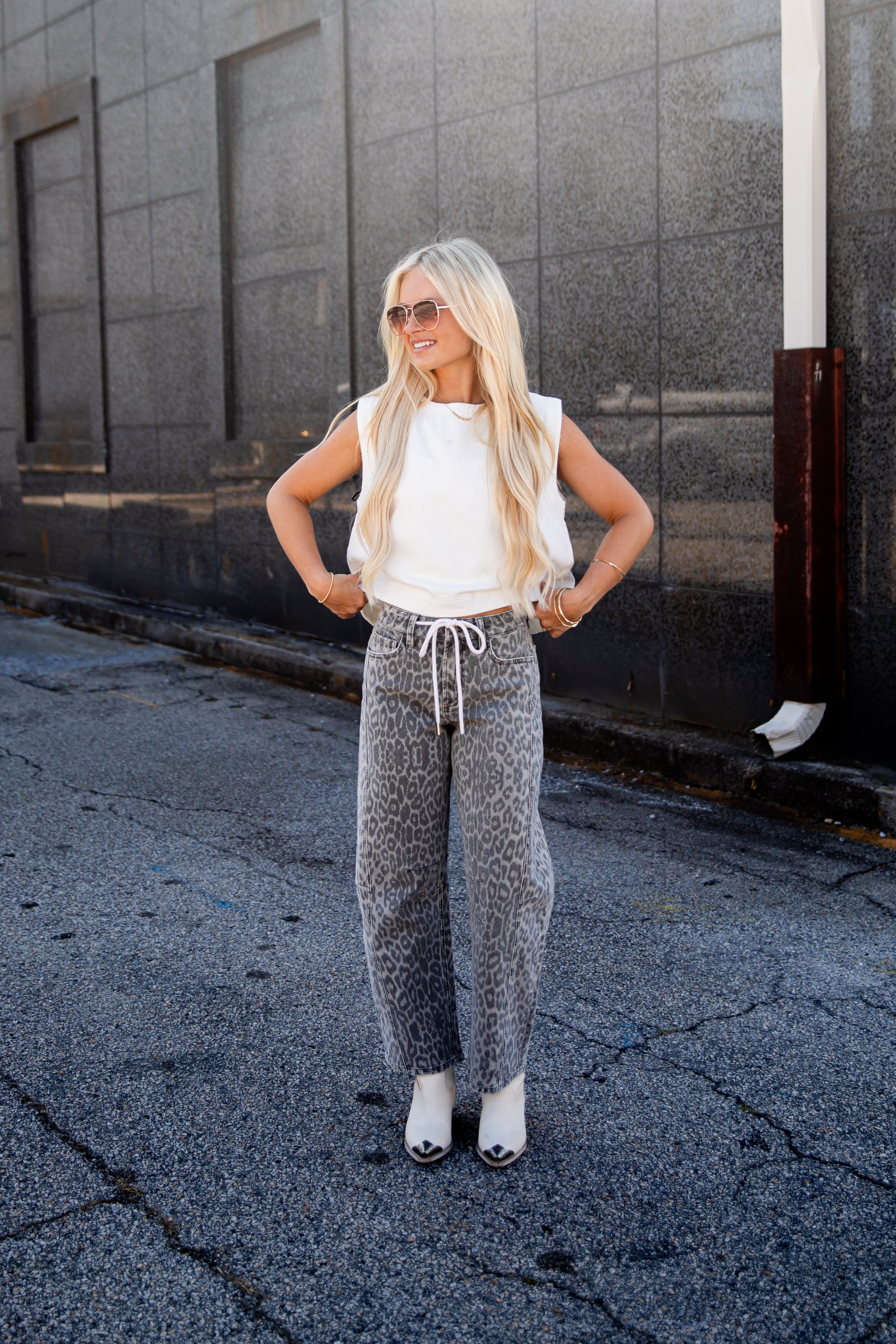 A woman stands outdoors on a paved street against a gray building wall, wearing gray leopard-print jogger sweatpants with a white drawstring, a white sleeveless top, white sneakers, and sunglasses.