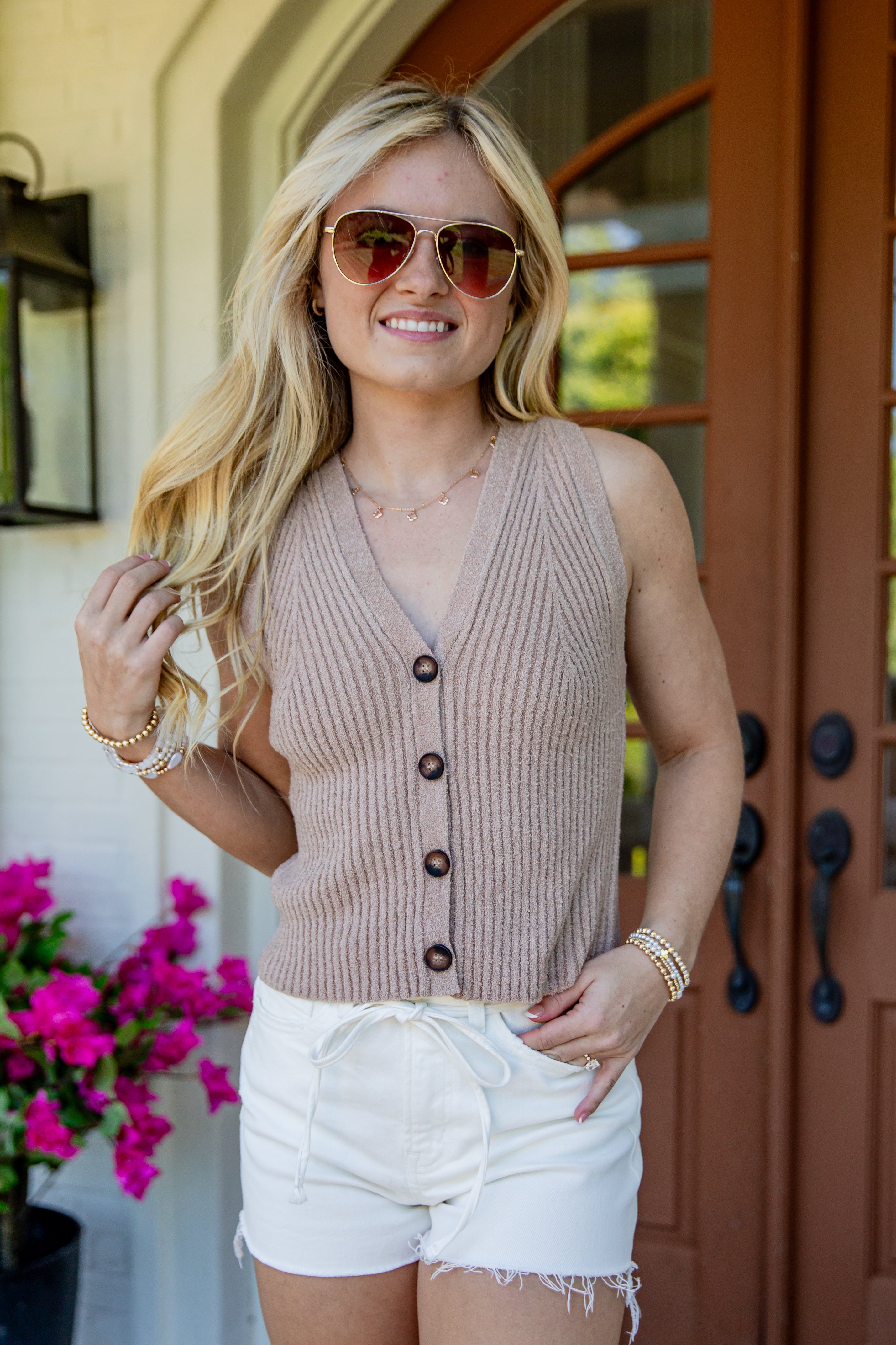 A woman wearing a sleeveless ribbed knit button-front vest in a warm taupe-beige color, paired with white shorts and sunglasses, standing in front of a brown wooden door.