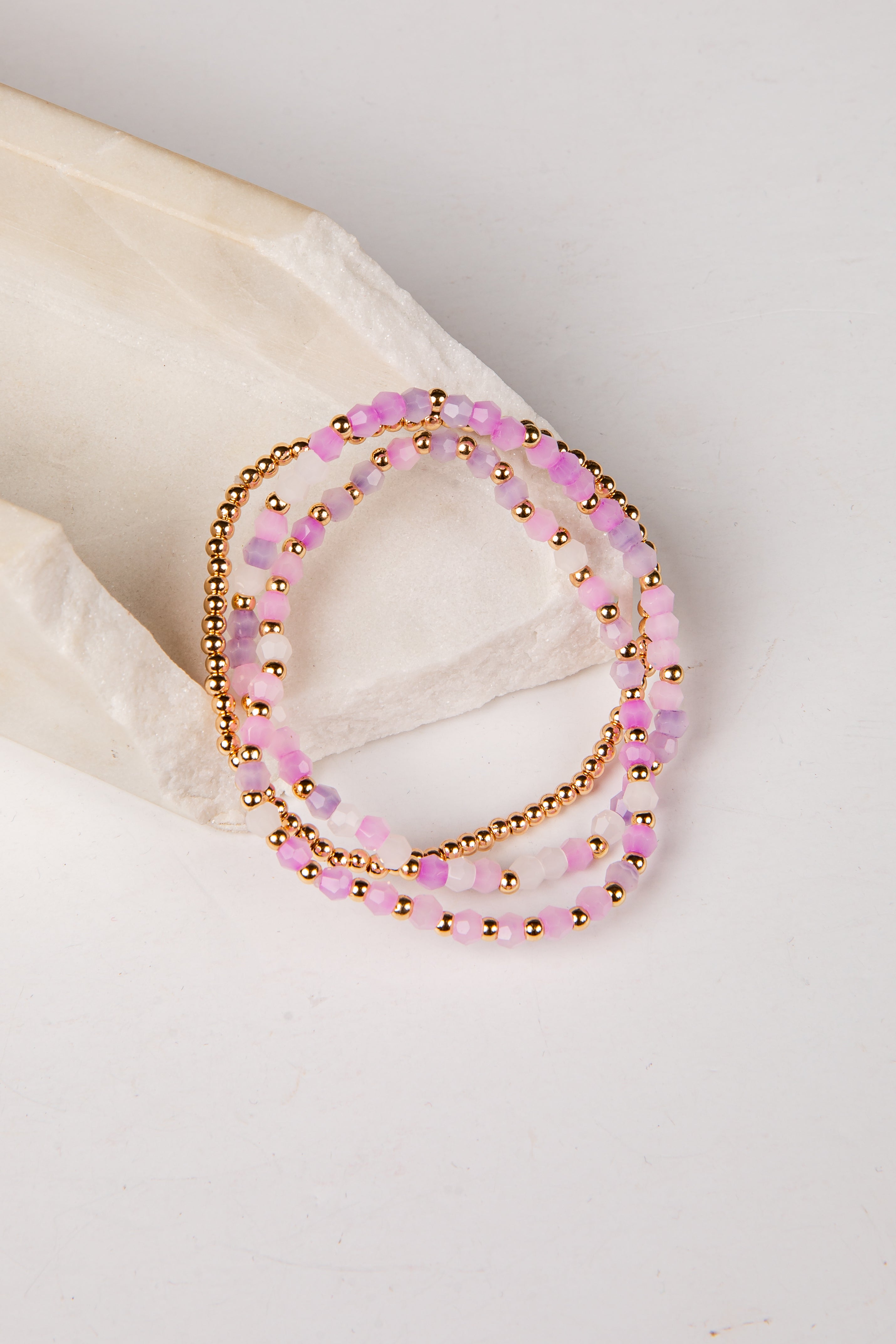 Three beaded stretch bracelets in pink and gold, displayed on a light beige textured stone prop against a white background.