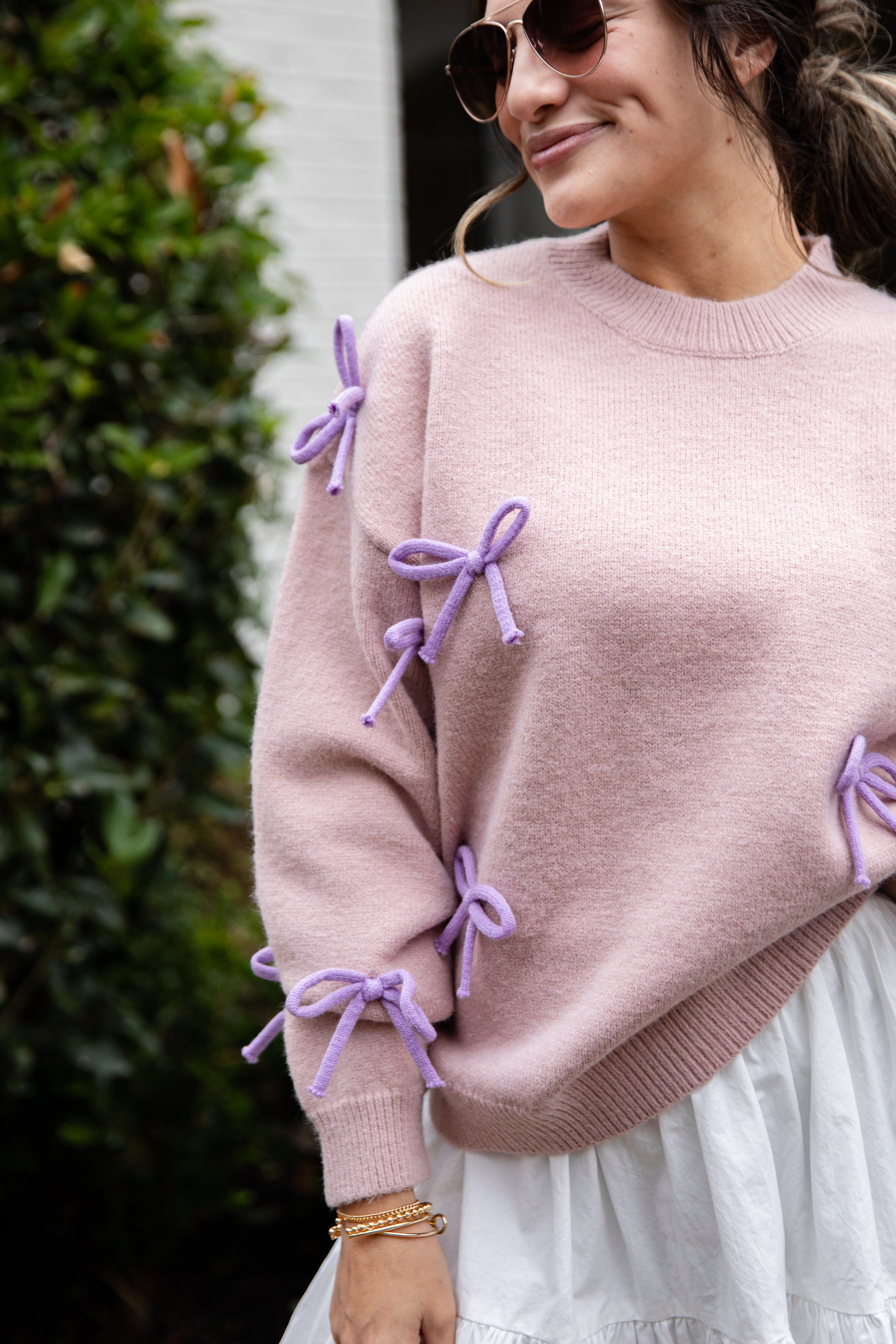 Woman wearing a pink sweater with purple bows and sunglasses outdoors.