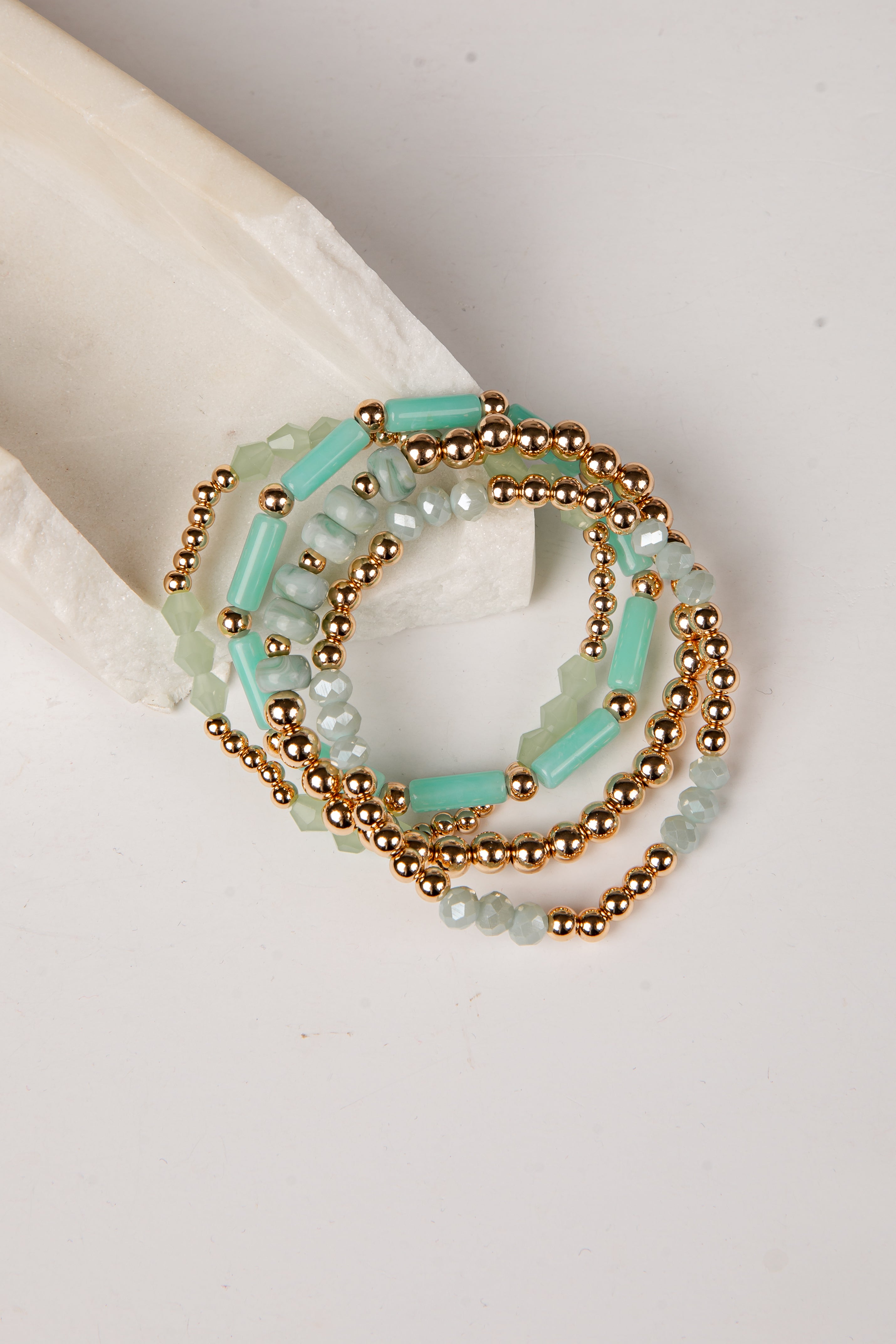 A stack of beaded stretch bracelets featuring light aqua green beads and metallic gold beads, displayed on a neutral cream-colored surface.