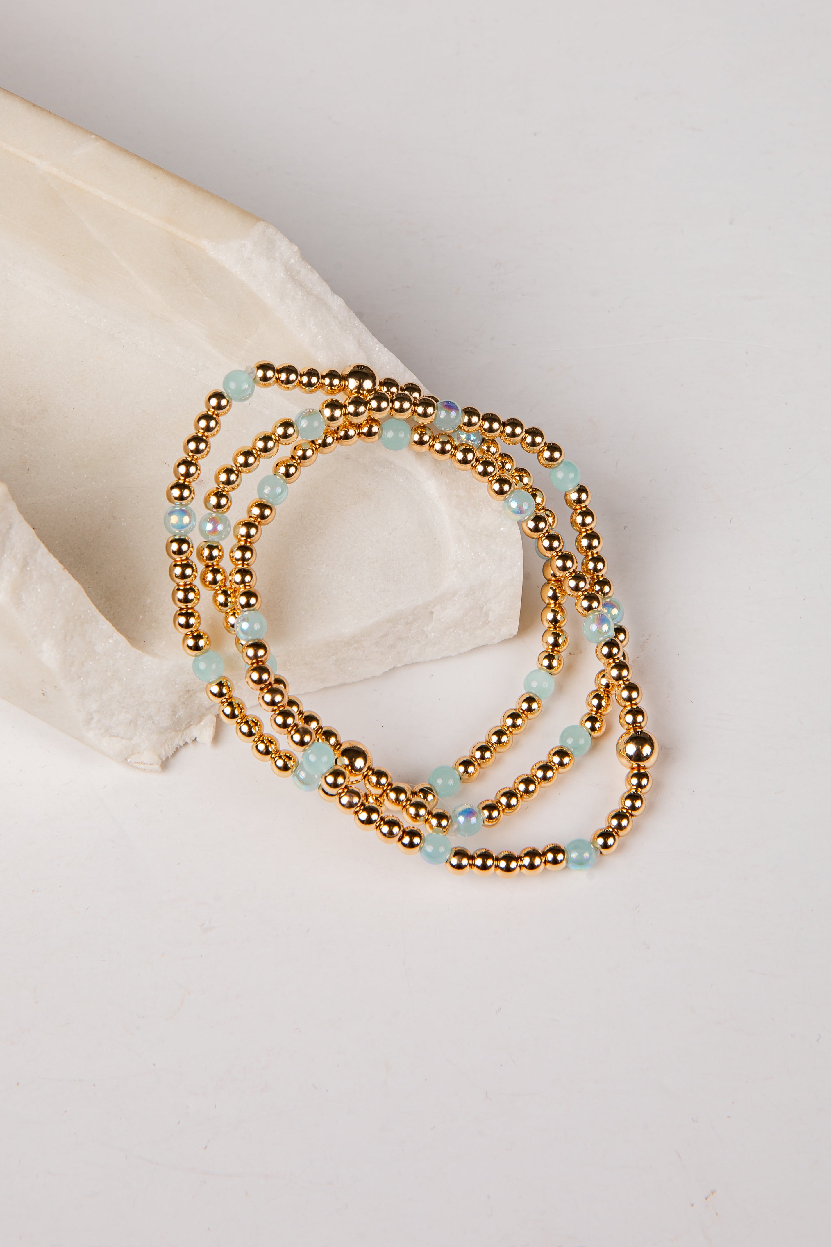 A multi-strand beaded necklace with metallic gold beads and light blue stone accents, styled on a white surface next to a cream-colored stone prop.