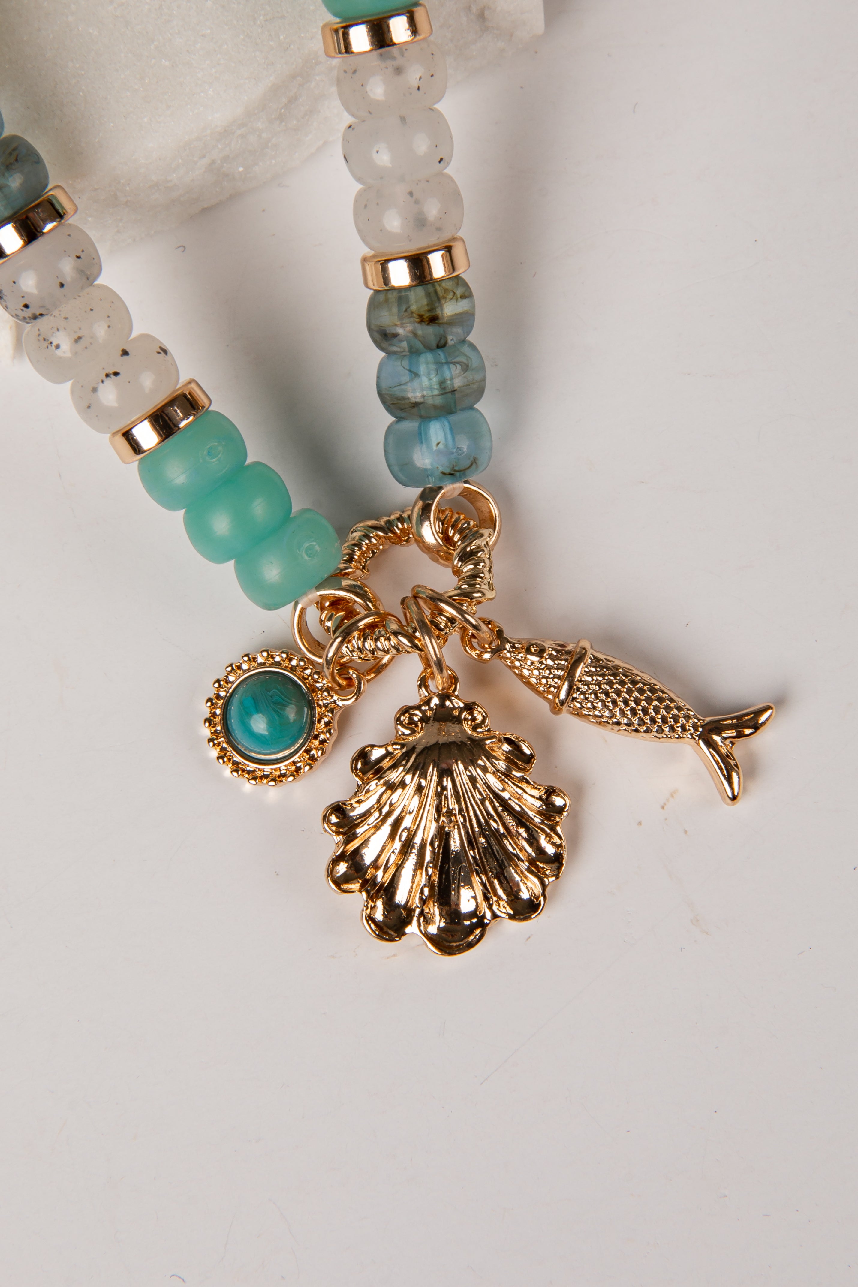 A close-up of a beaded jewelry charm strand with gold-tone metal charms, including a seashell, fish, and turquoise stone, alongside aqua and marbled beads on a white background.