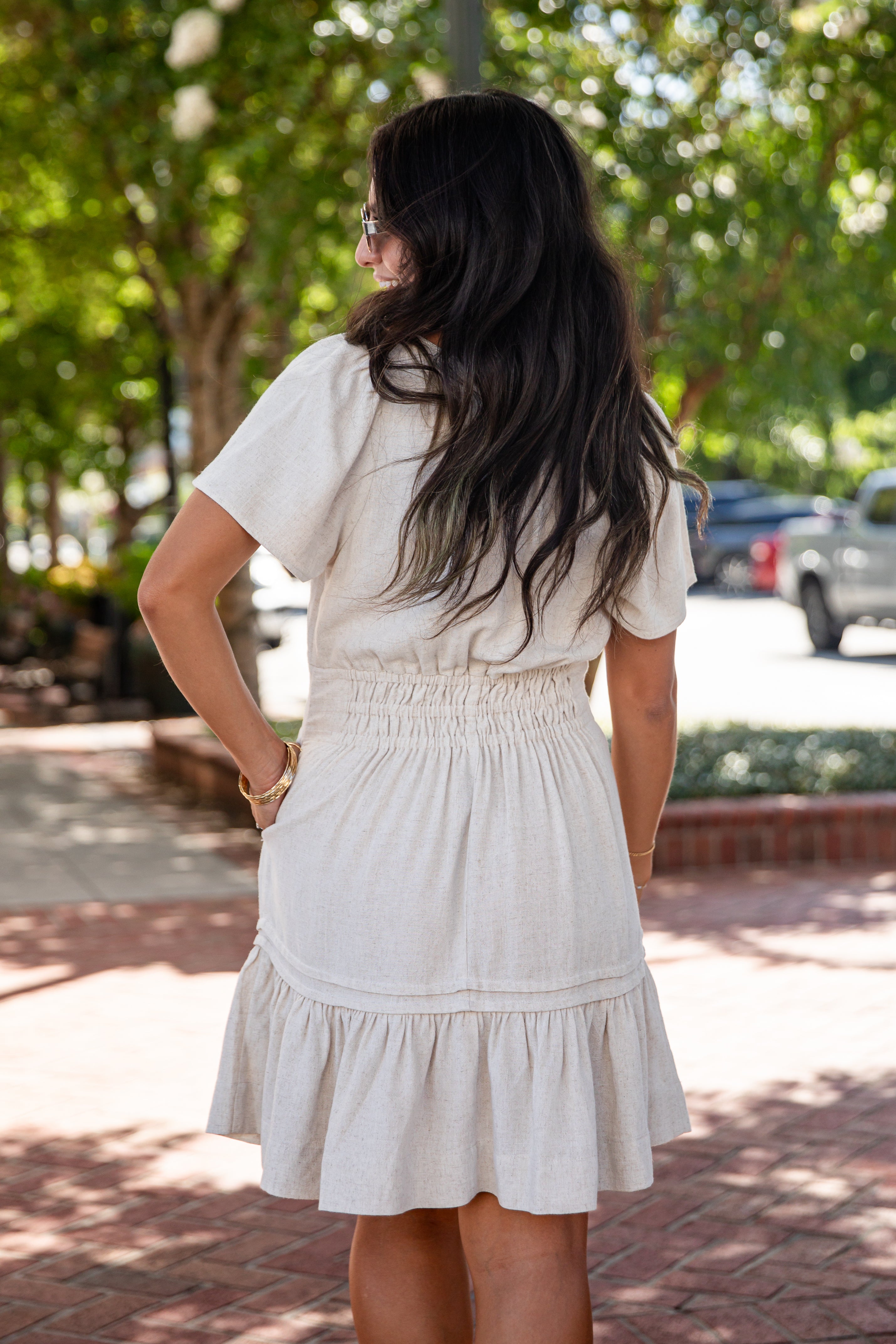 A woman viewed from behind wearing a short-sleeve cream-colored dress with a smocked waist and tiered skirt, standing outdoors on a brick walkway with trees and parked cars in the background.