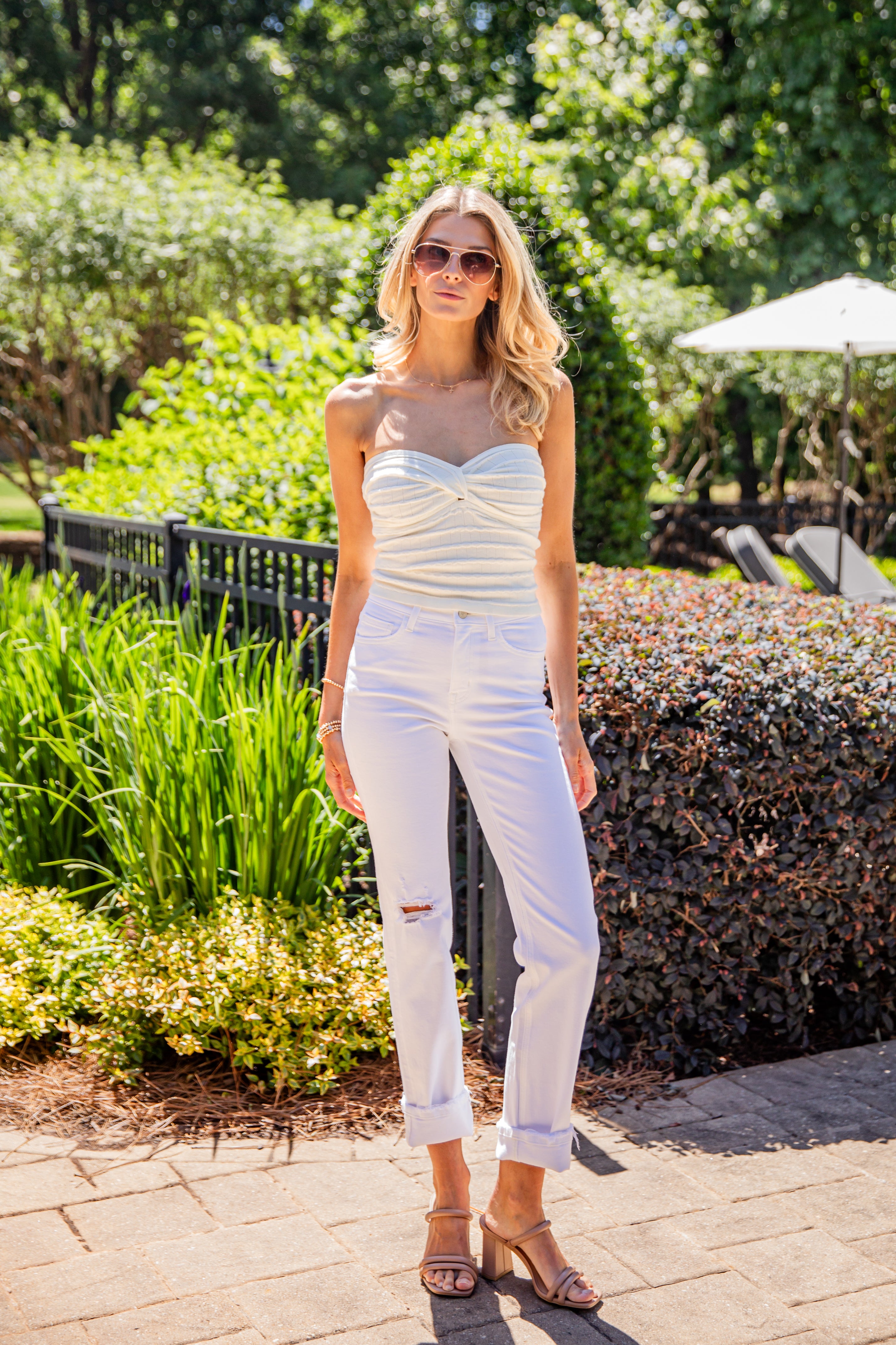 Woman in a strapless top and white pants standing outdoors with greenery in the background