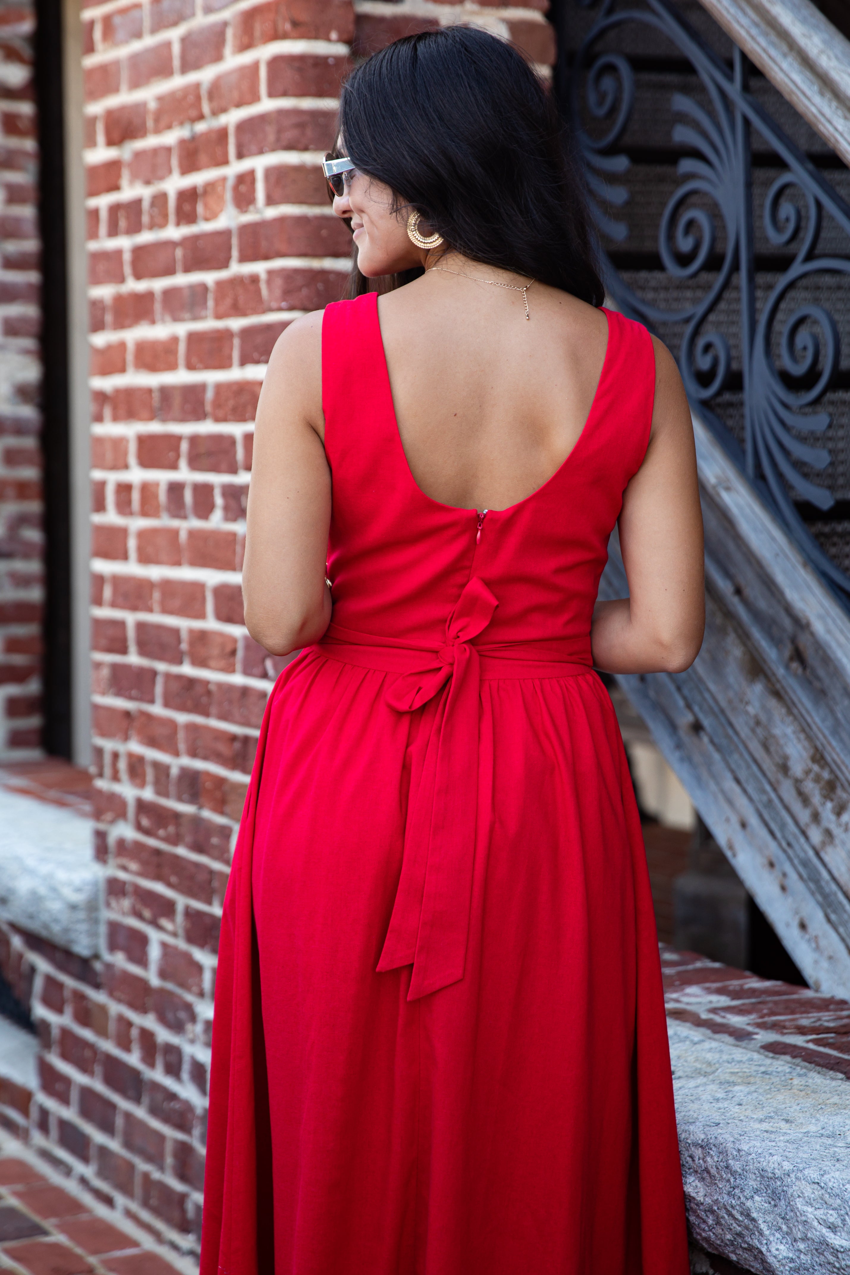 A woman wearing a sleeveless bright red maxi dress with a deep V-neckline and a tied waist bow, photographed from behind in an outdoor setting next to a brick wall and metal staircase.