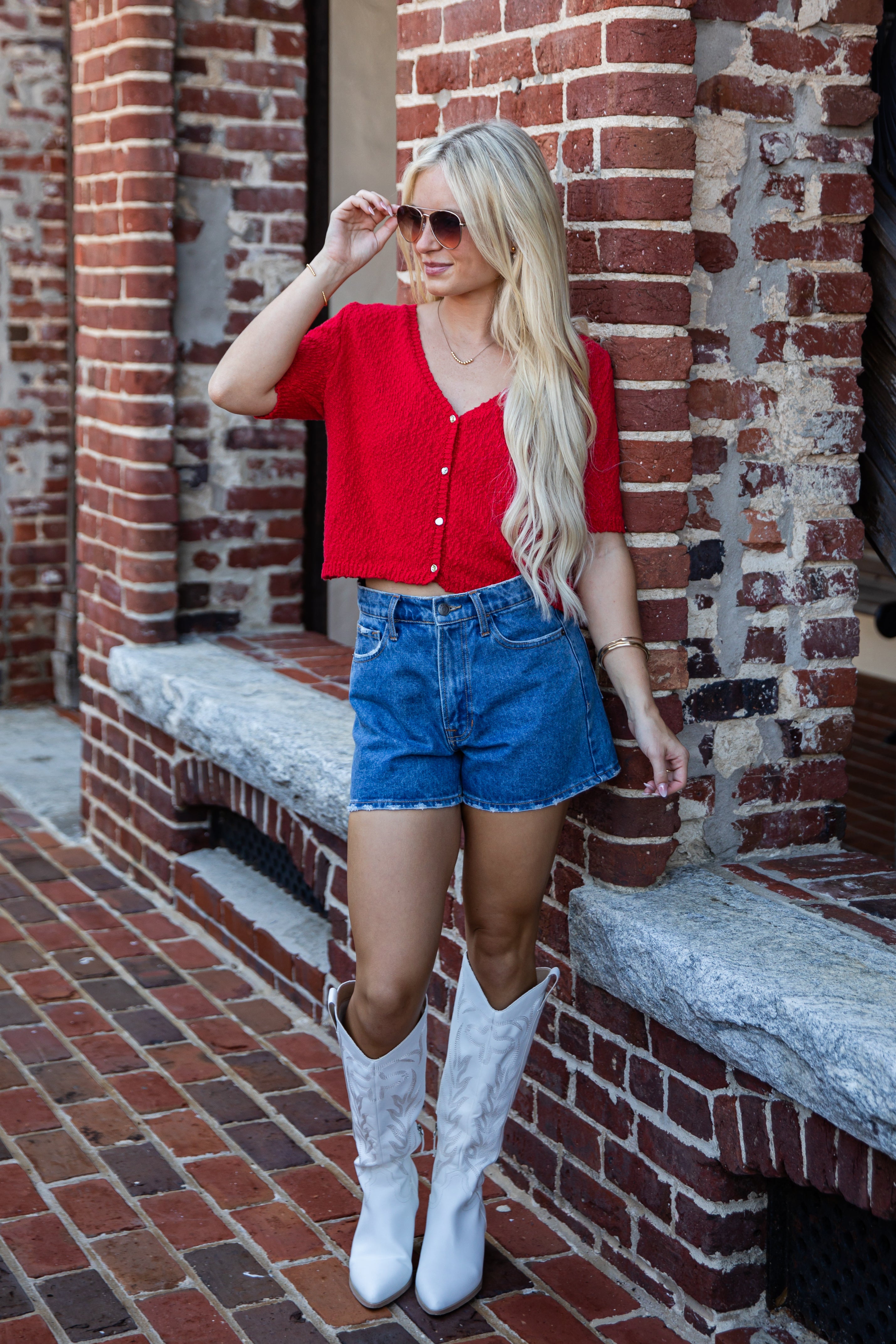 A woman stands outdoors against a red brick building, wearing a bright red short-sleeve button-front top, blue denim shorts, and white cowboy boots.