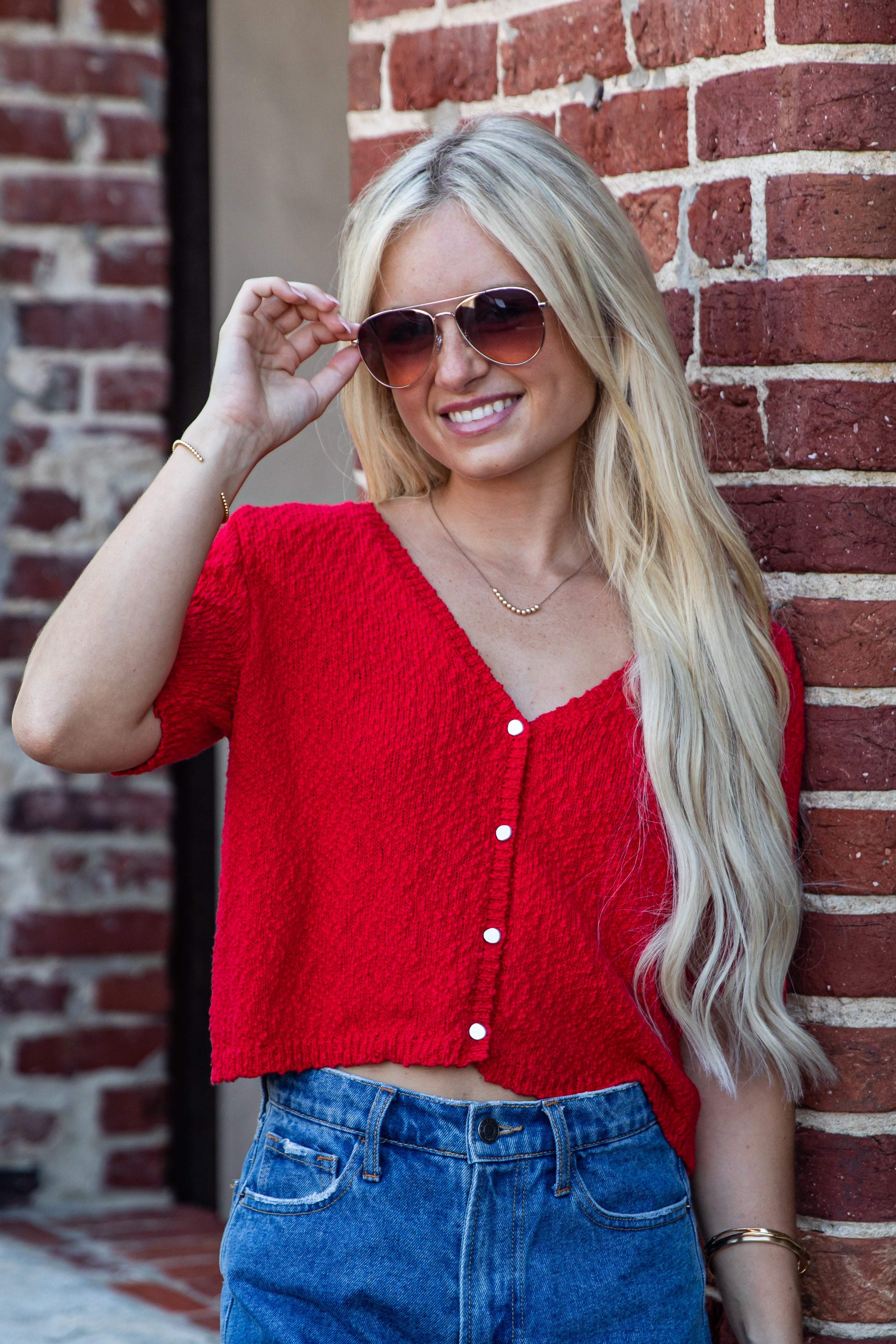 A woman with long blonde hair wearing a bright red textured short-sleeve crop top and blue jeans, posing against a red brick wall.