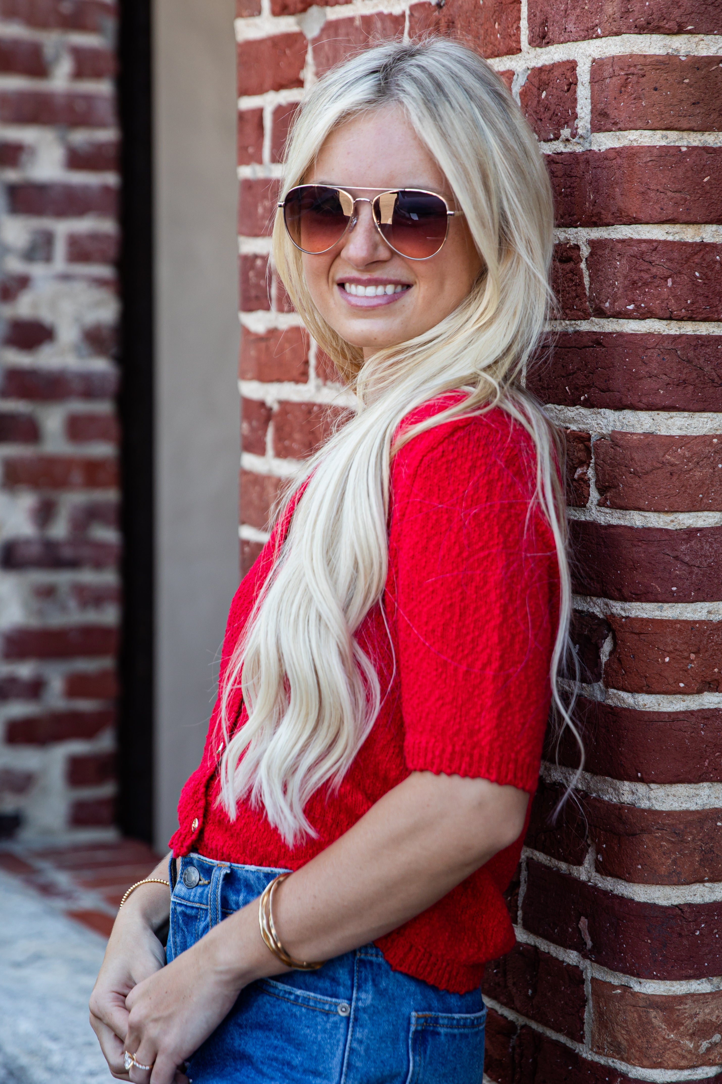 A smiling blonde woman wearing aviator sunglasses, a bright red knit short-sleeve top, and blue jeans, posing against a red brick wall.