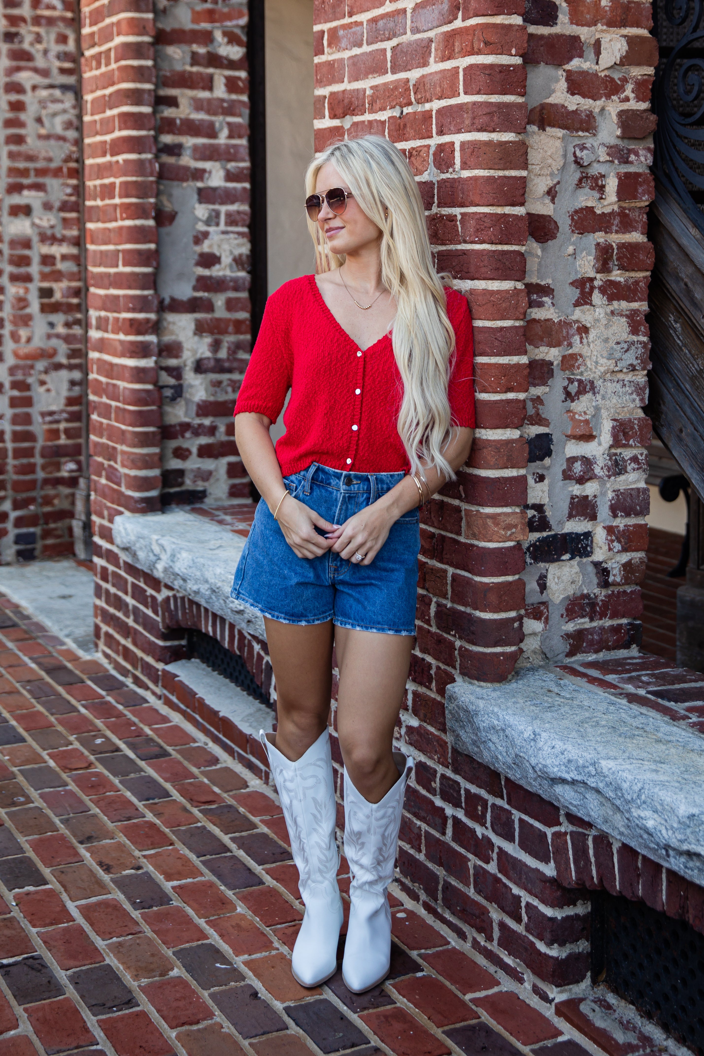 A woman stands against a red brick building wearing a bright red short-sleeve button-front top, blue denim shorts, and white cowboy boots.