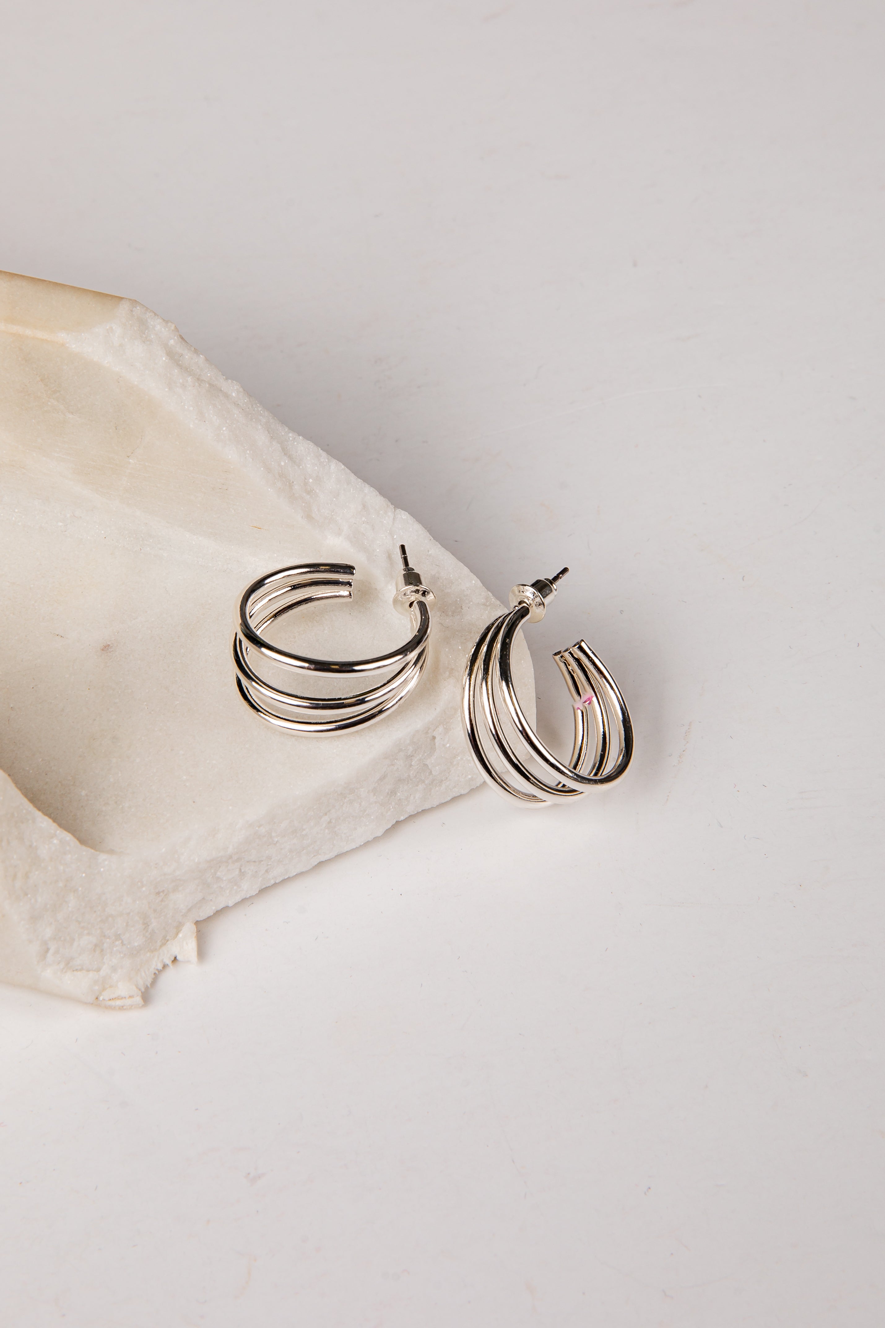A pair of silver-toned multi-band hoop earrings resting on a textured beige stone slab against a clean white background.