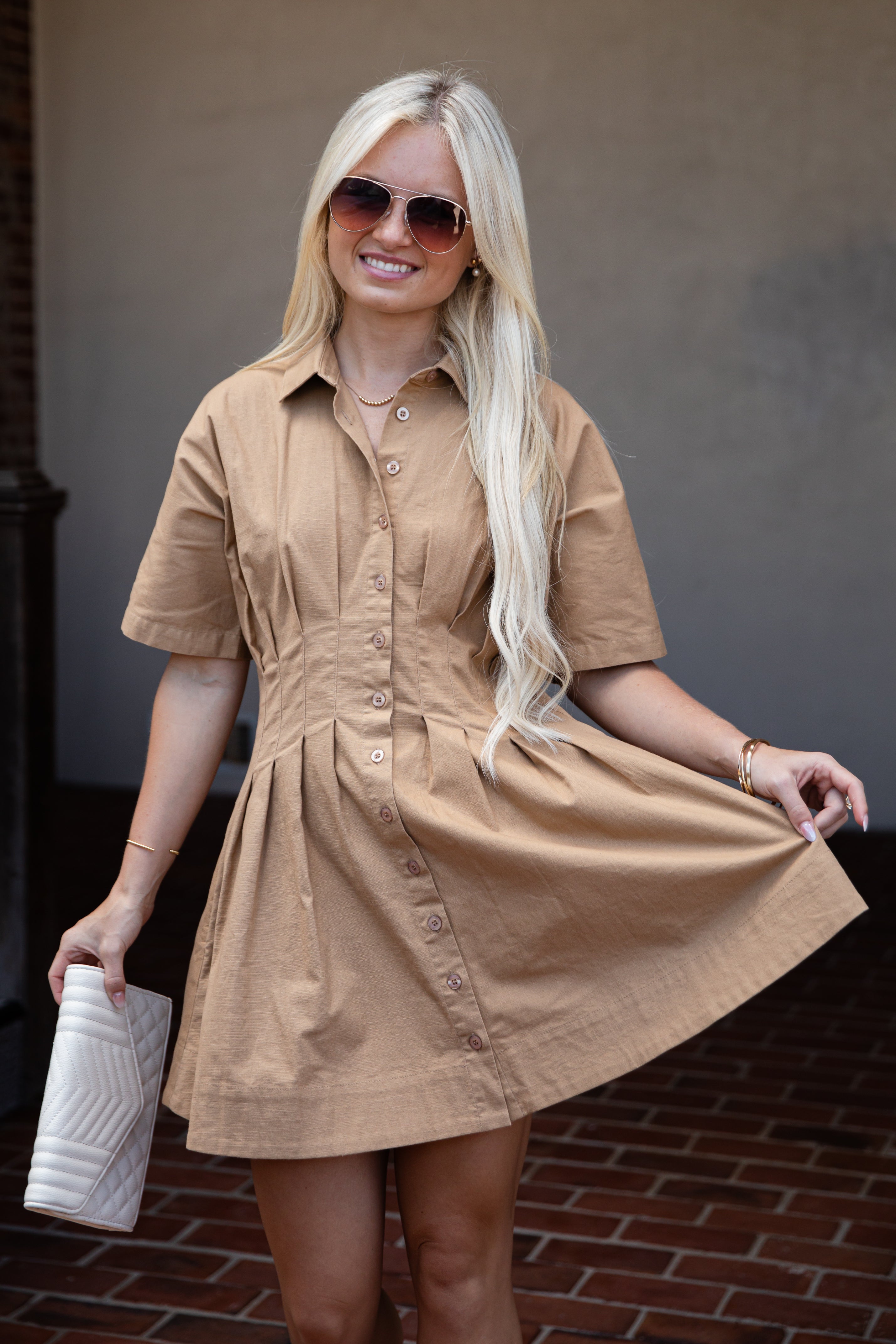A blonde woman wearing a tan short-sleeve button-front shirt dress and sunglasses, holding out the skirt of the dress to show its shape, while holding a white clutch in one hand.