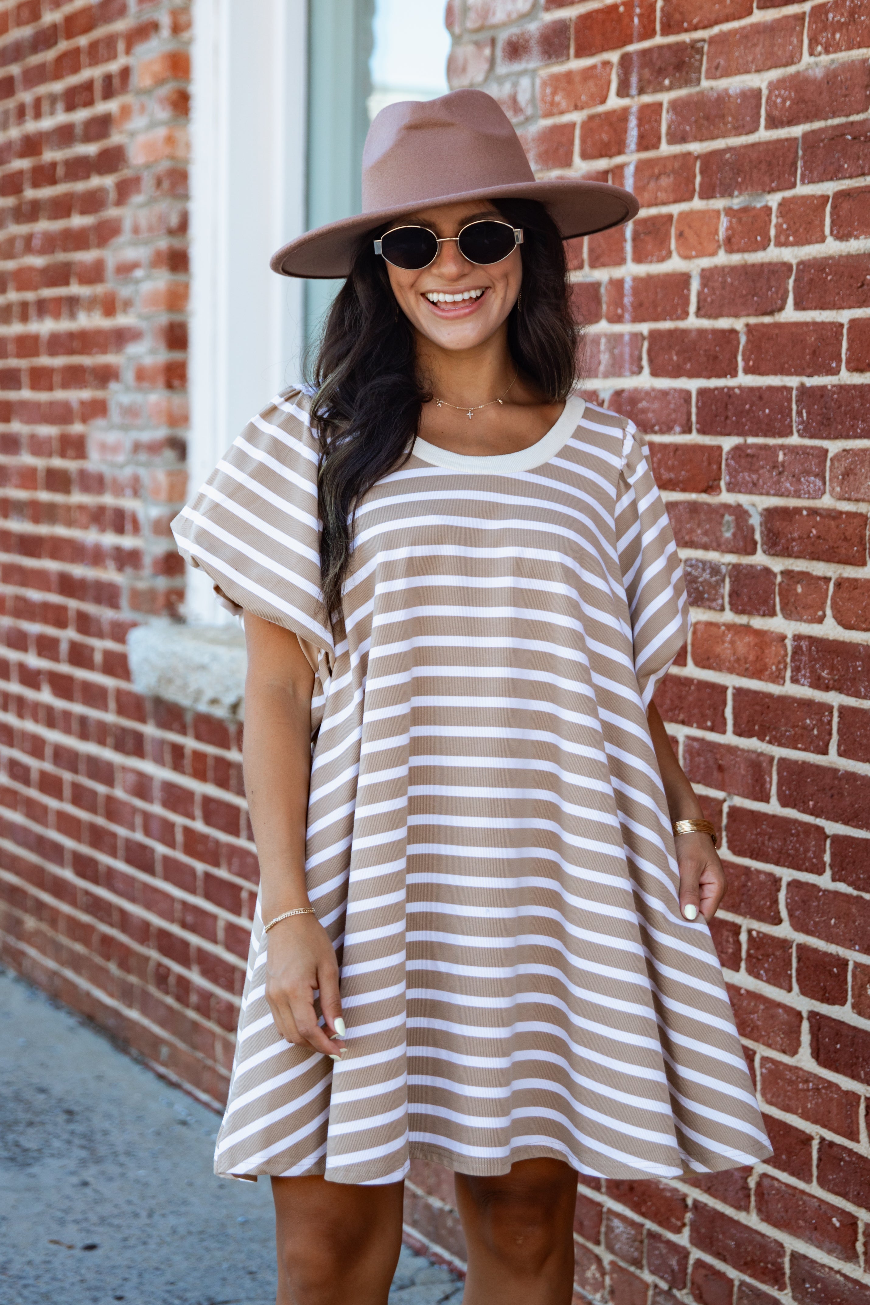 A woman wearing a brown wide-brim hat, sunglasses, and a beige and white striped tunic dress stands in front of a red brick wall.