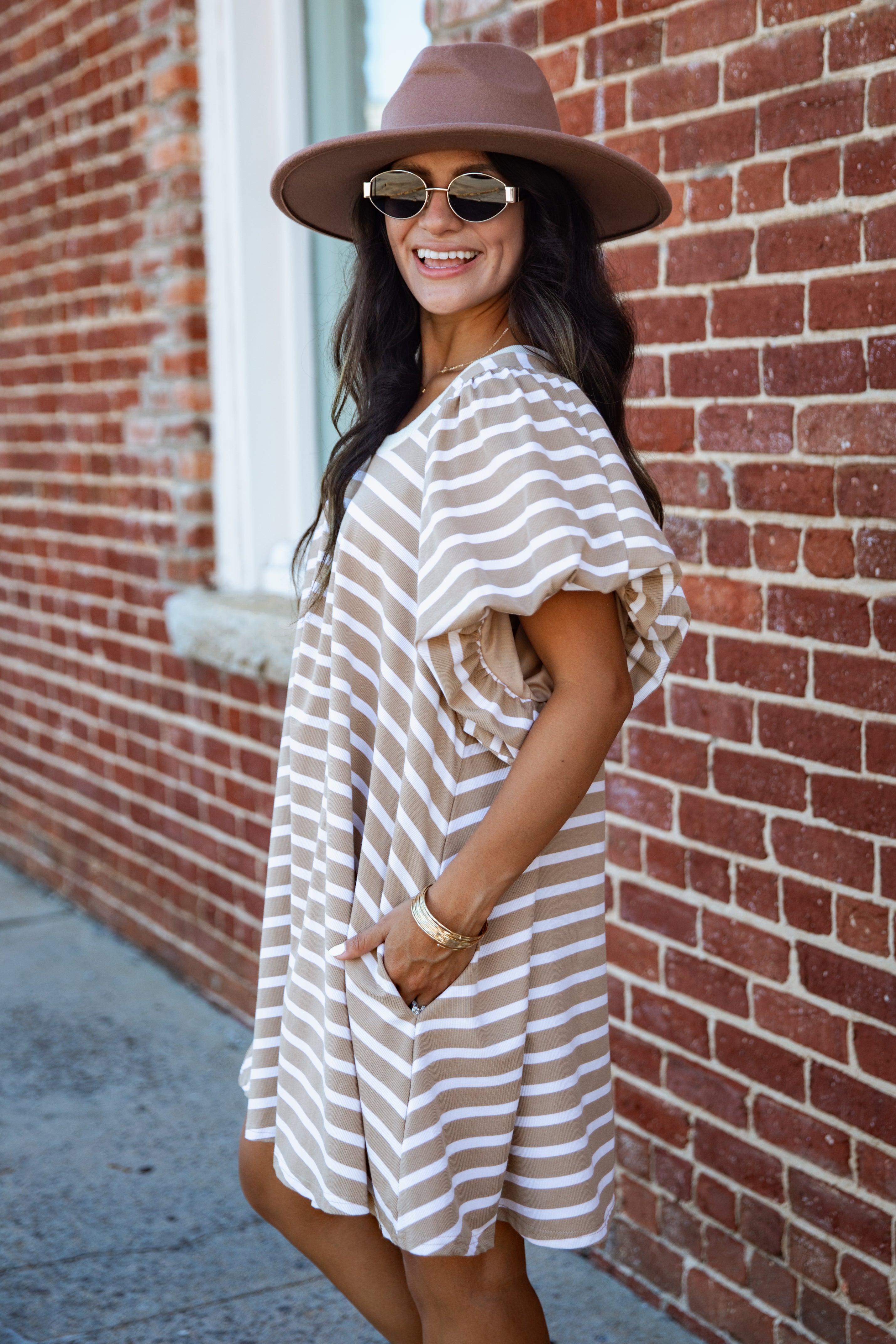 A woman wearing a brown wide-brim hat, sunglasses, and a beige and white striped short dress stands in front of a red brick wall.