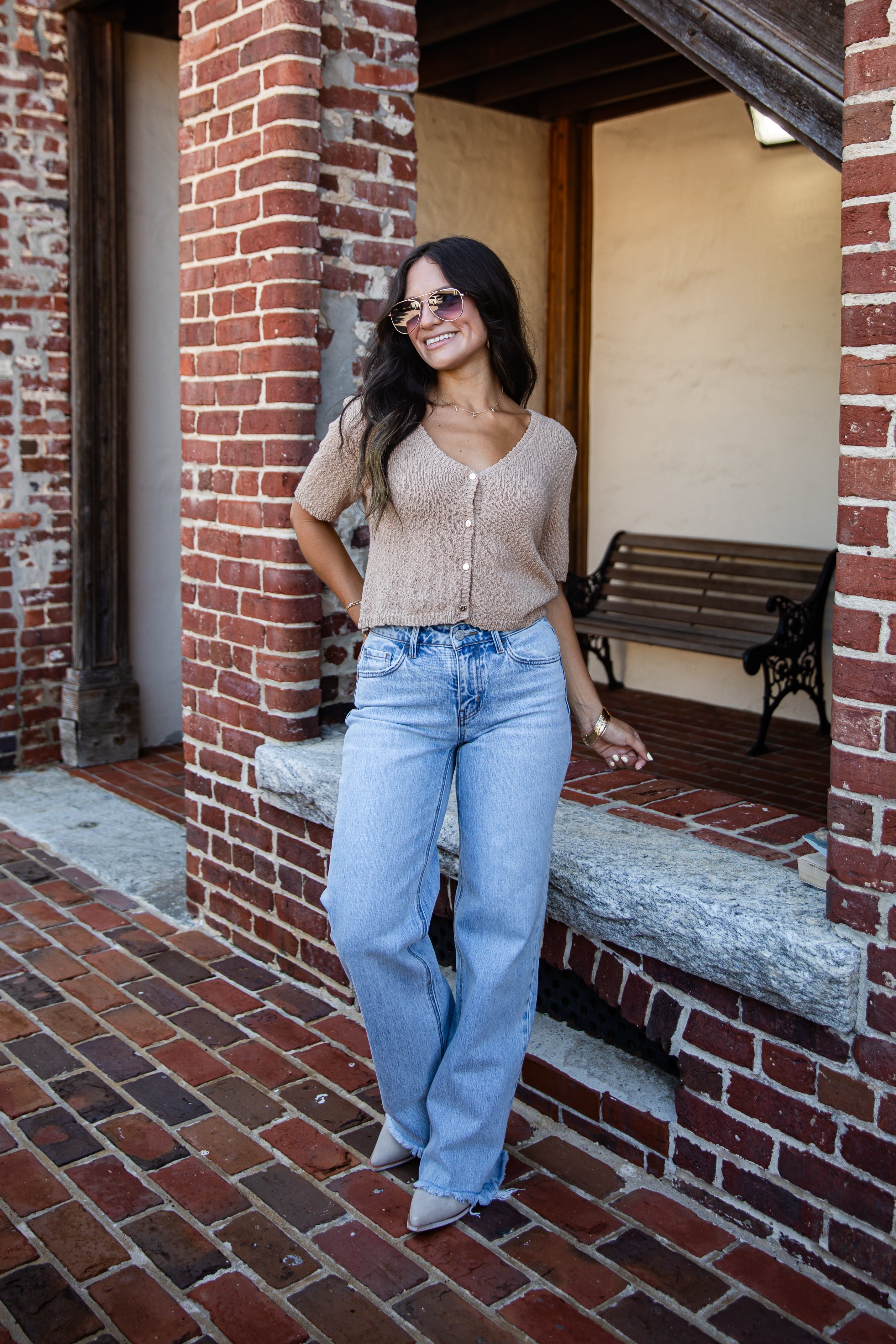 A woman wearing a beige knit V-neck top and light blue wide-leg jeans stands in a brick walkway next to a building with a wooden bench in the background.