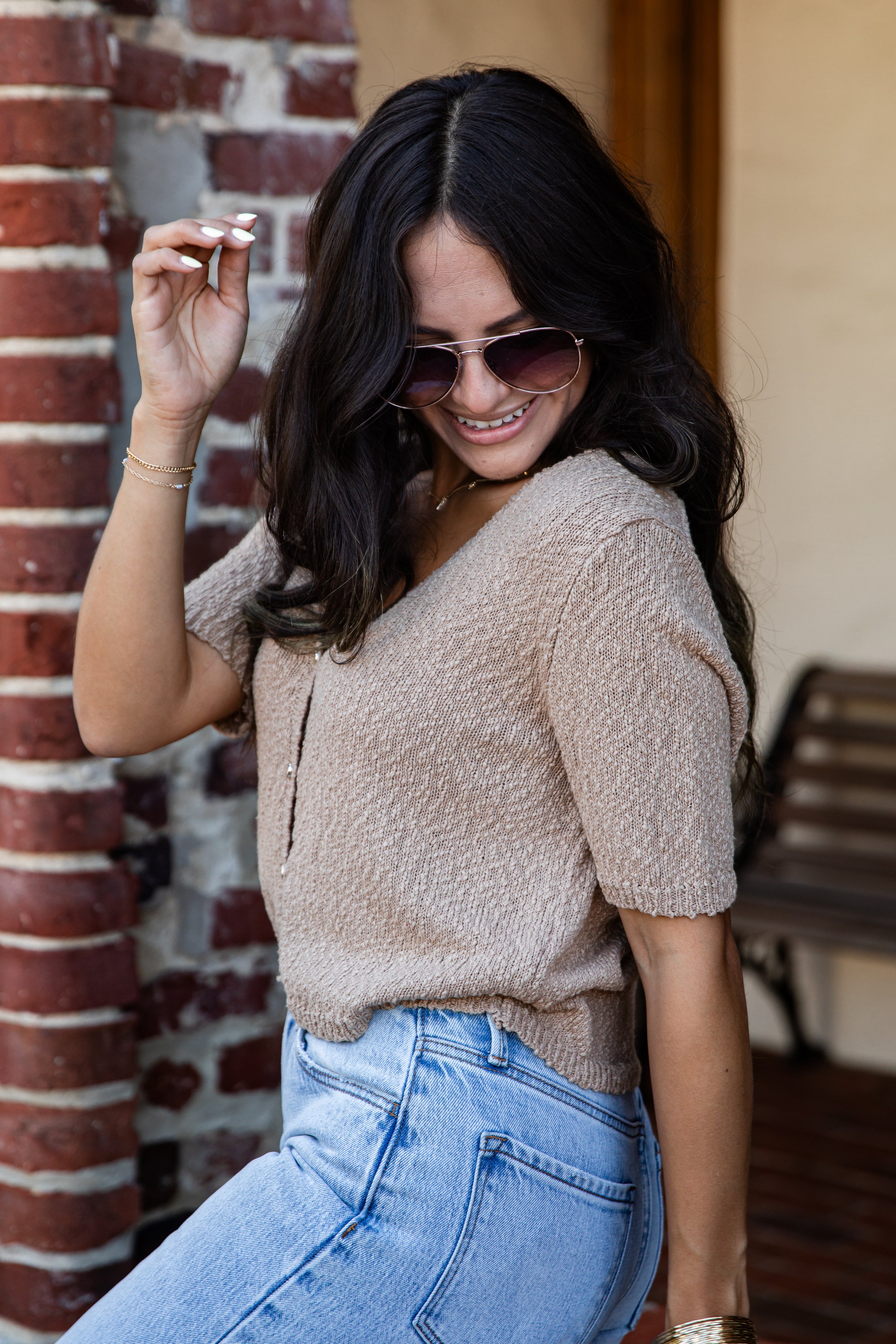 A woman wearing a beige short-sleeve knit top and light blue jeans, posing outdoors next to a brick column.