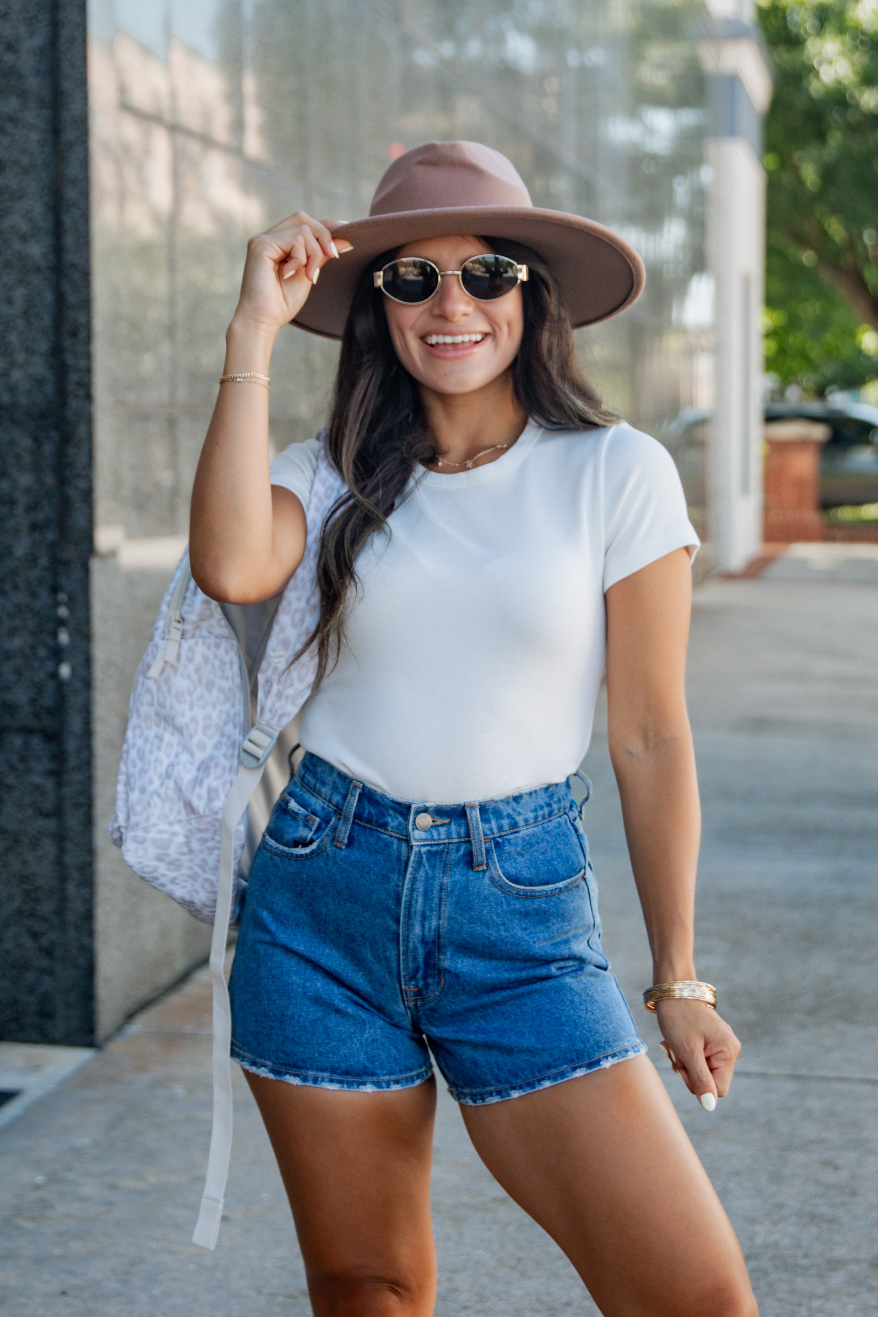 A woman posing outdoors in a white t-shirt, blue denim shorts, a brown wide-brim hat, and sunglasses, carrying a light blue patterned bag.