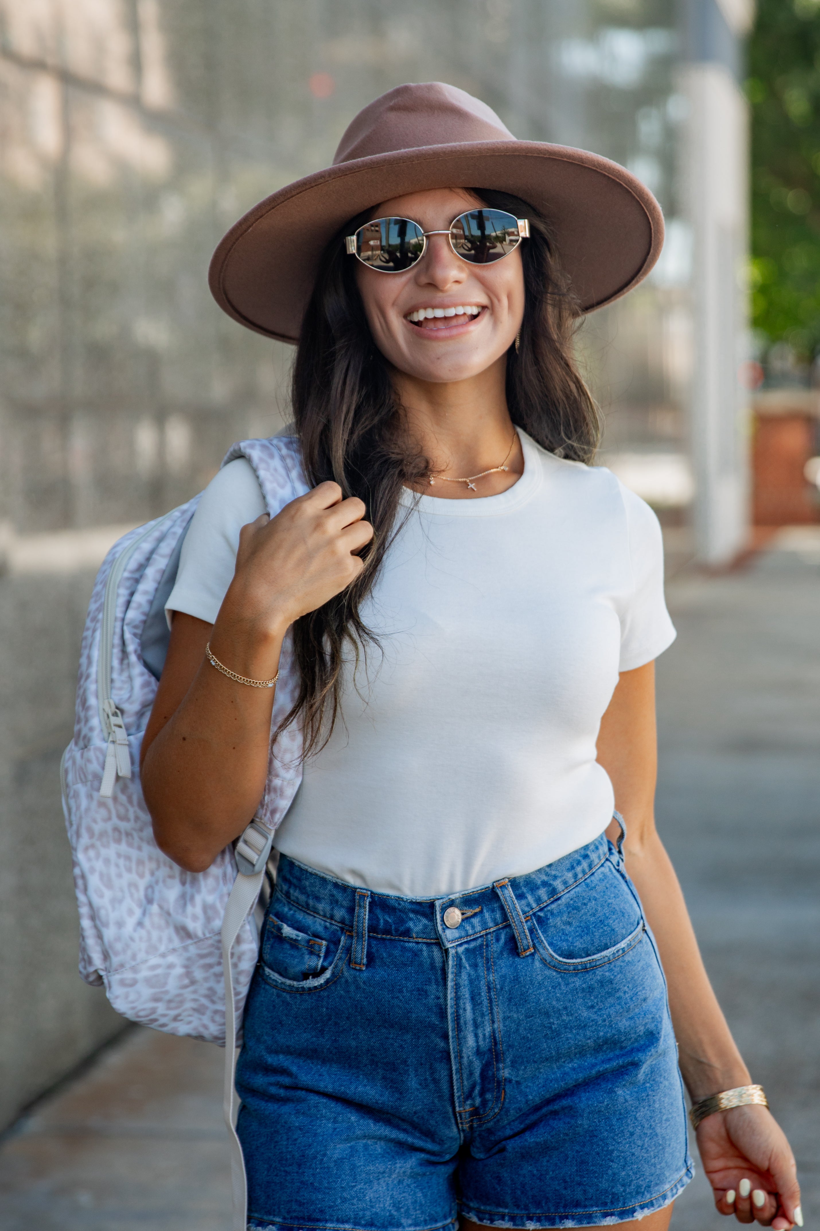 A woman wearing a brown wide-brim hat, round sunglasses, a white t-shirt, and blue denim shorts, carrying a light patterned backpack on her shoulder in an outdoor urban setting.