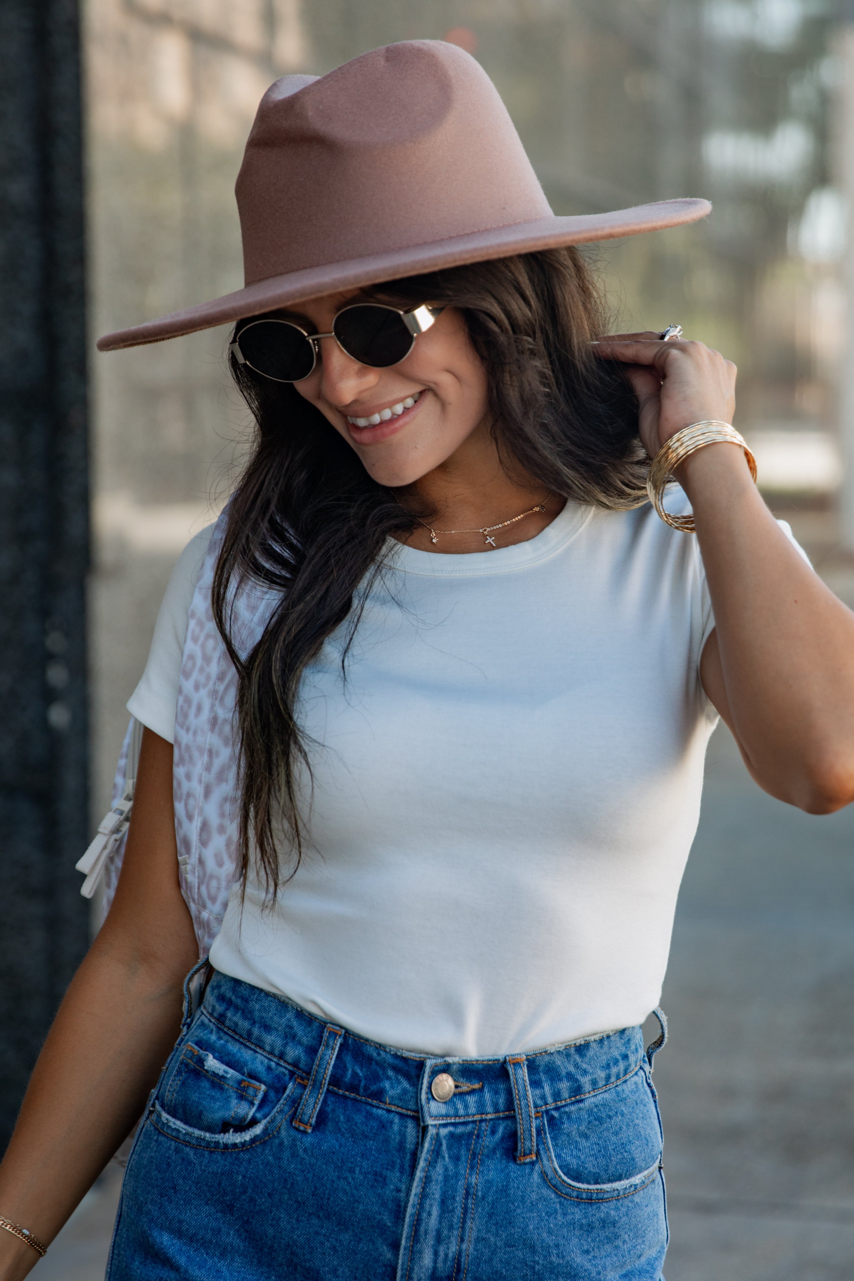 A woman wearing a dusty pink wide-brim fedora hat, round black sunglasses, a white t-shirt, and blue jeans, posing outdoors.