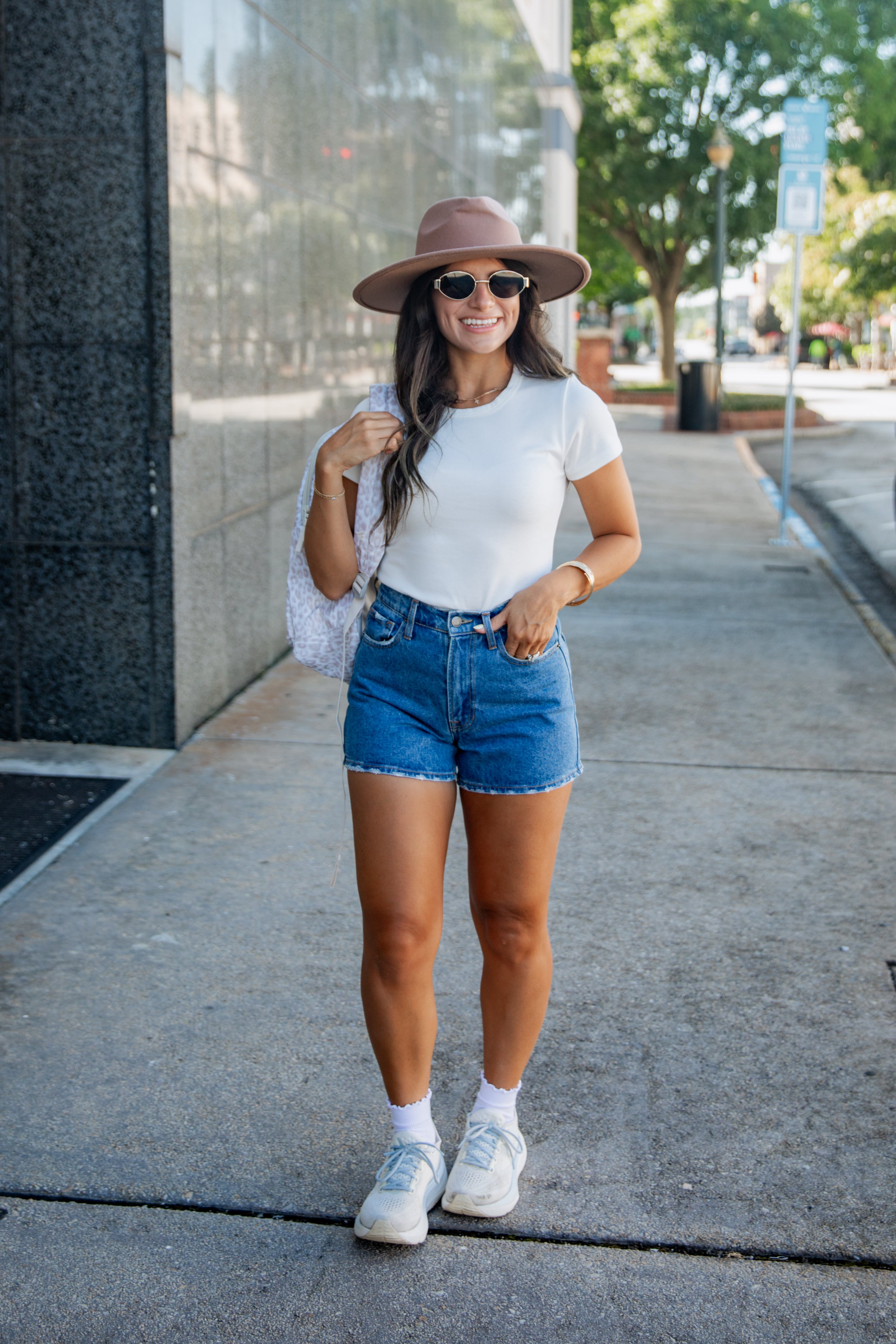 A woman stands on a city sidewalk wearing a white short-sleeve T-shirt, blue denim cutoff shorts, a brown wide-brim hat, white sneakers, and carrying a white shoulder bag.