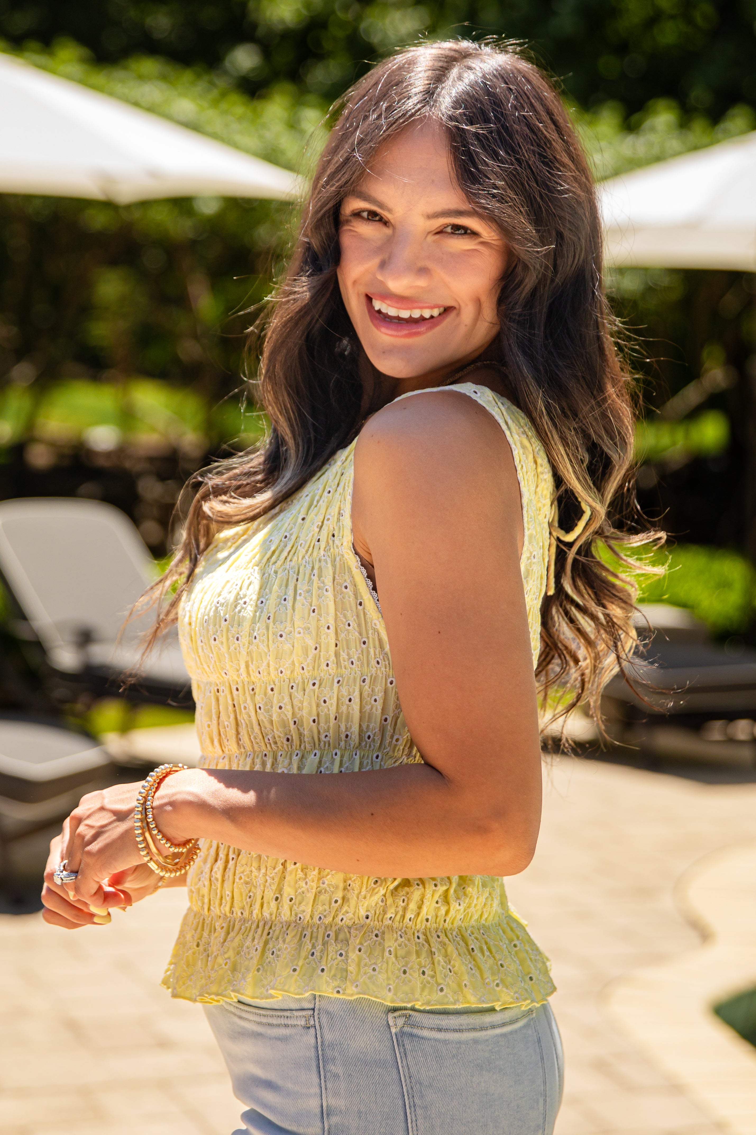 Woman in a yellow top and blue jeans standing outdoors with greenery in the background