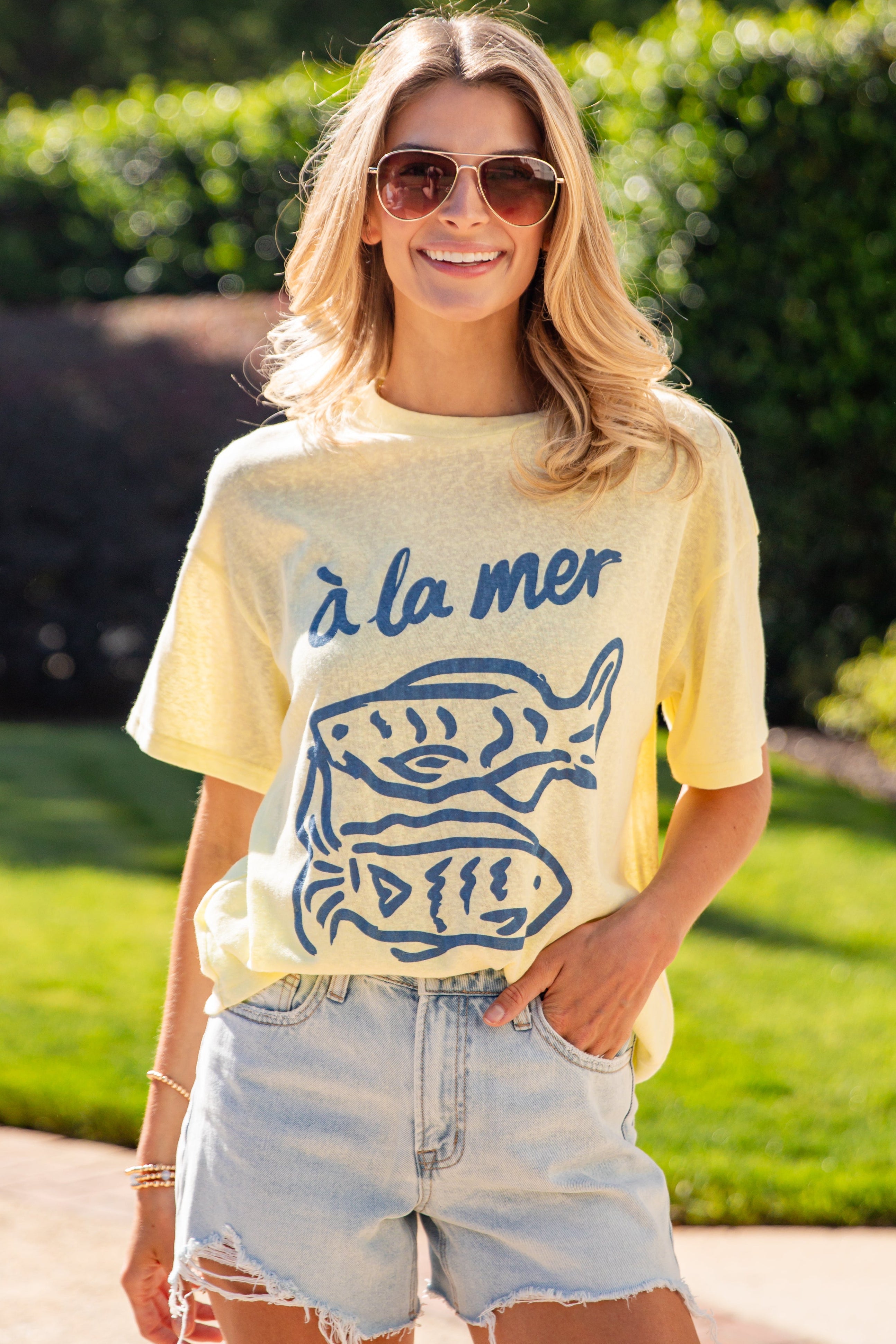 Woman wearing a yellow t-shirt with blue graphics and text, standing outdoors.