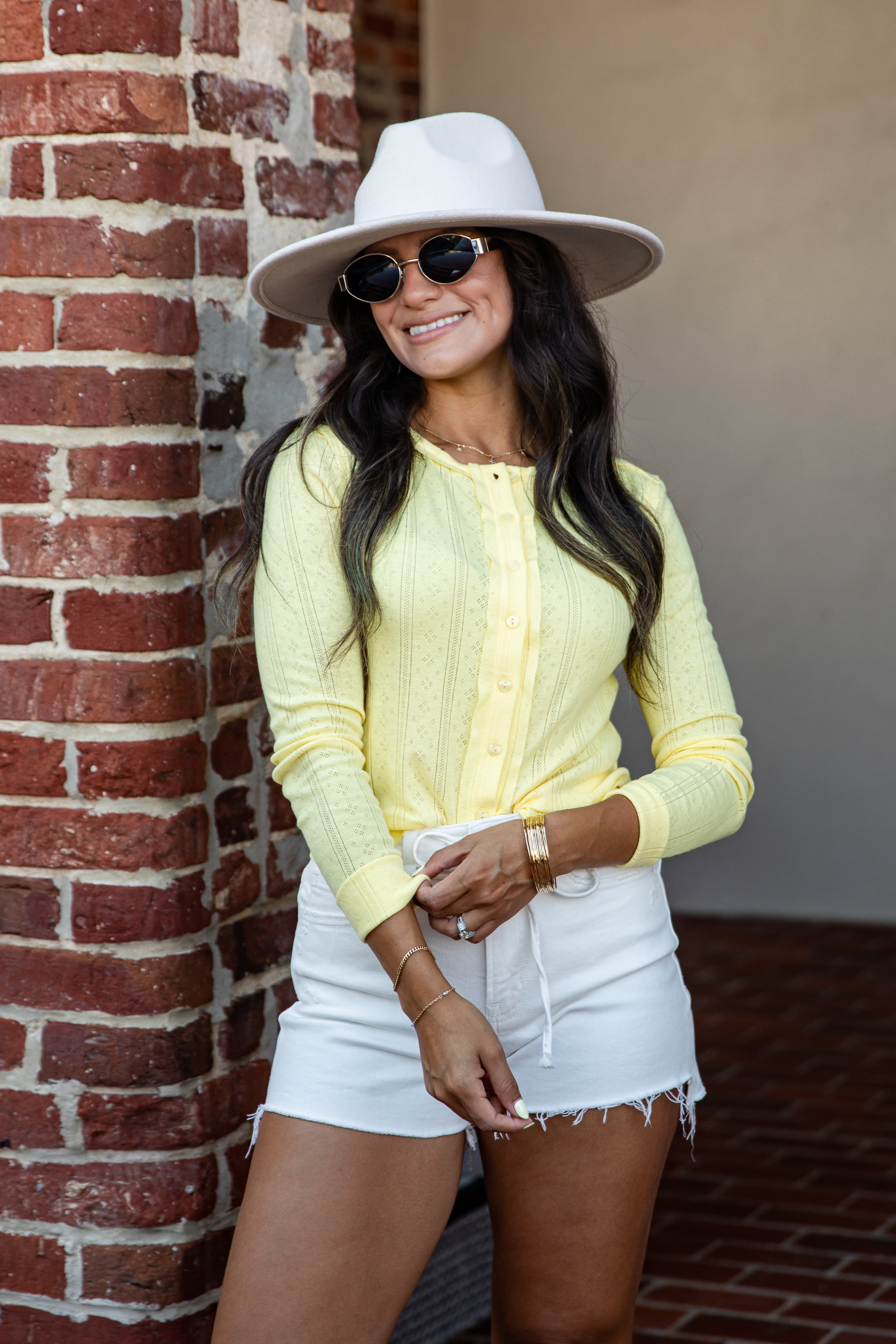 A woman wearing a pale yellow ribbed knit long-sleeve top, white denim shorts, a white wide-brim hat, and sunglasses, posing next to a red brick column.