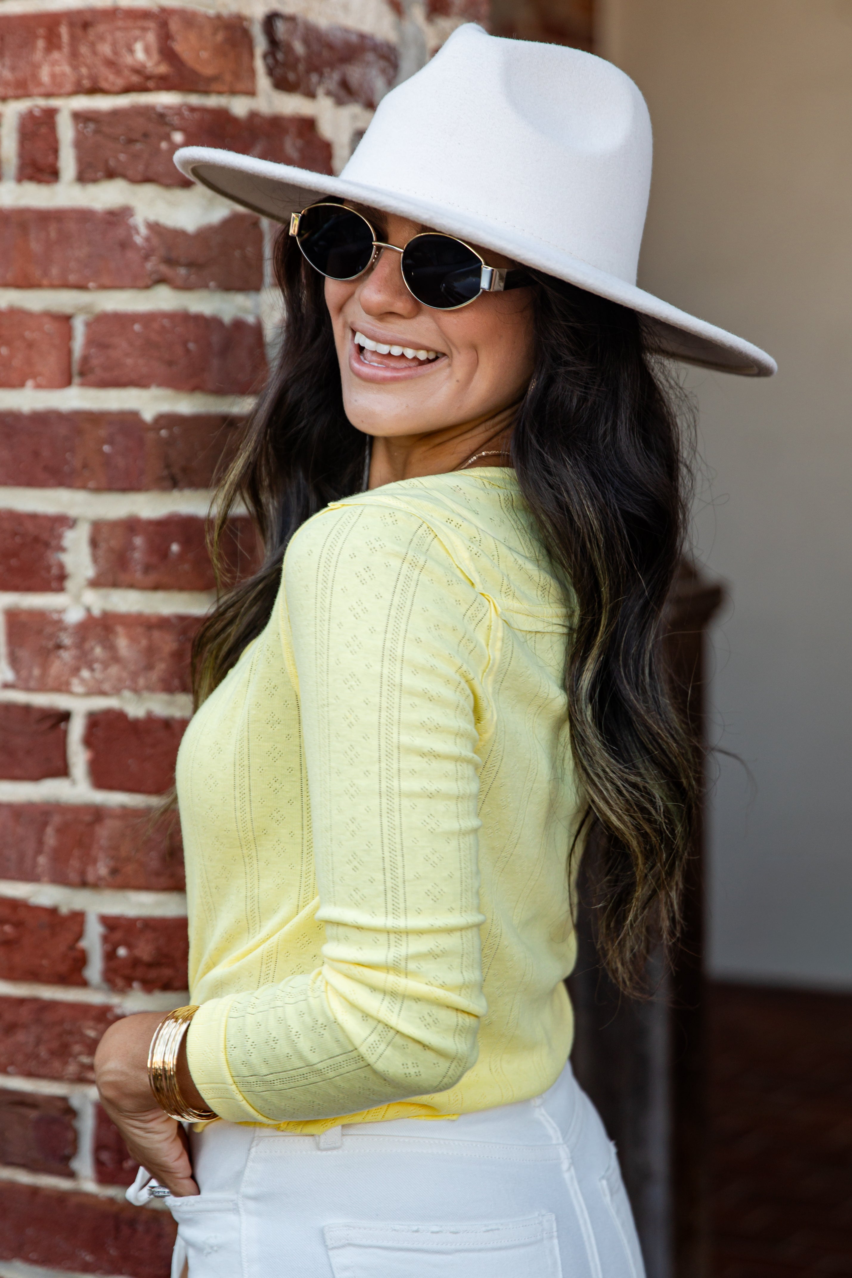 A woman wearing a white wide-brim fedora hat, round sunglasses, a pale yellow ribbed knit long-sleeve top, and white pants, posing in front of a red brick wall.