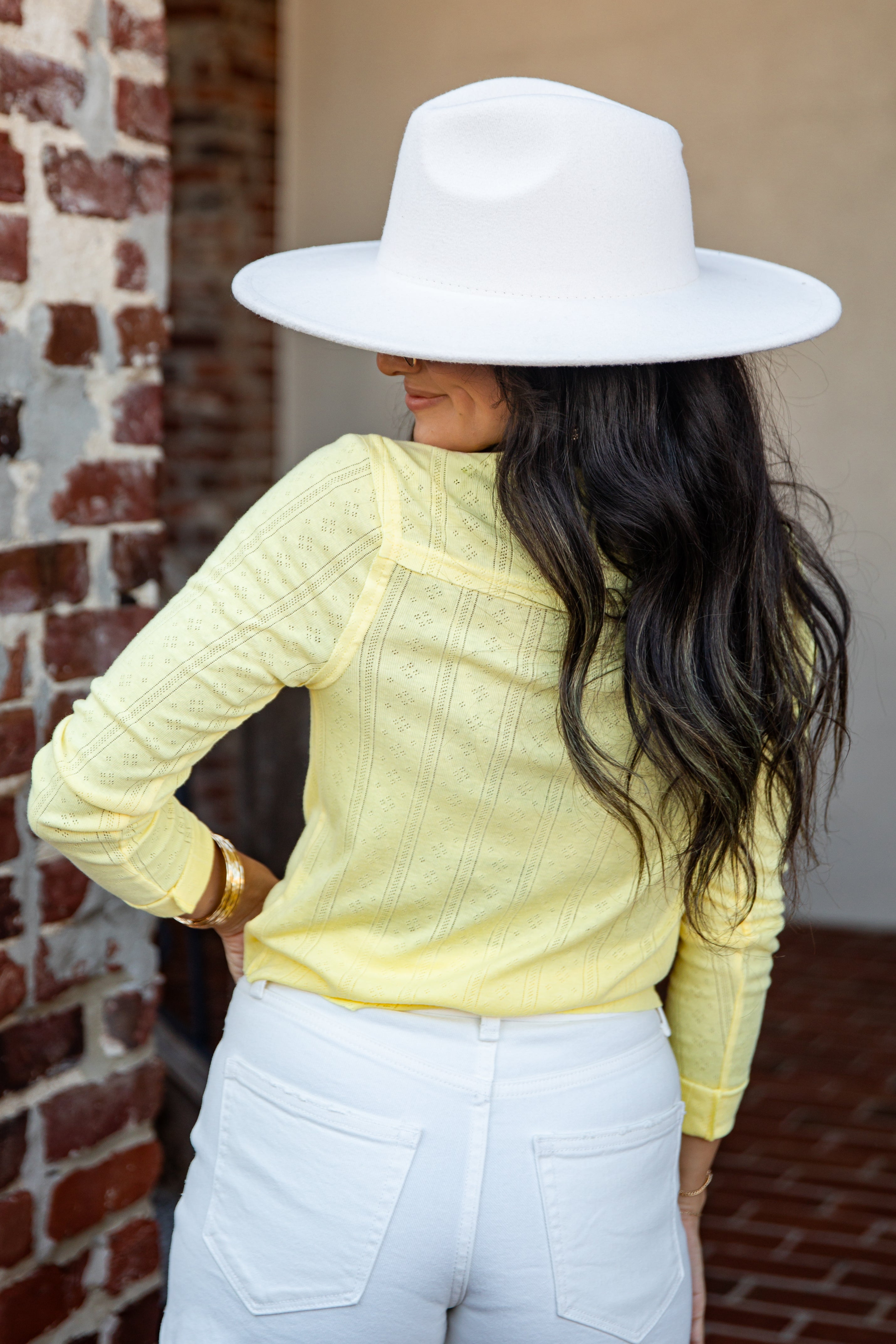 A woman shown from the back wearing a pale yellow textured knit top, white pants, and a large white wide-brim hat, standing outdoors next to a brick wall.
