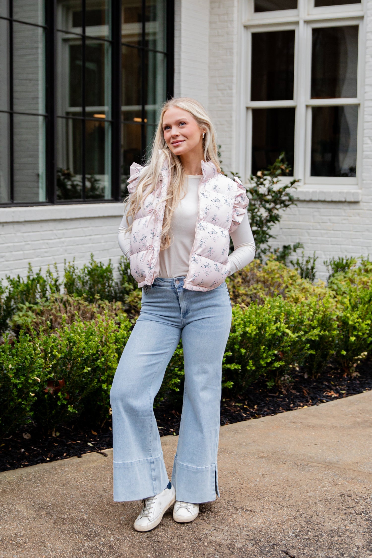 Woman standing outside wearing a pink puffer vest and jeans with white sneakers
