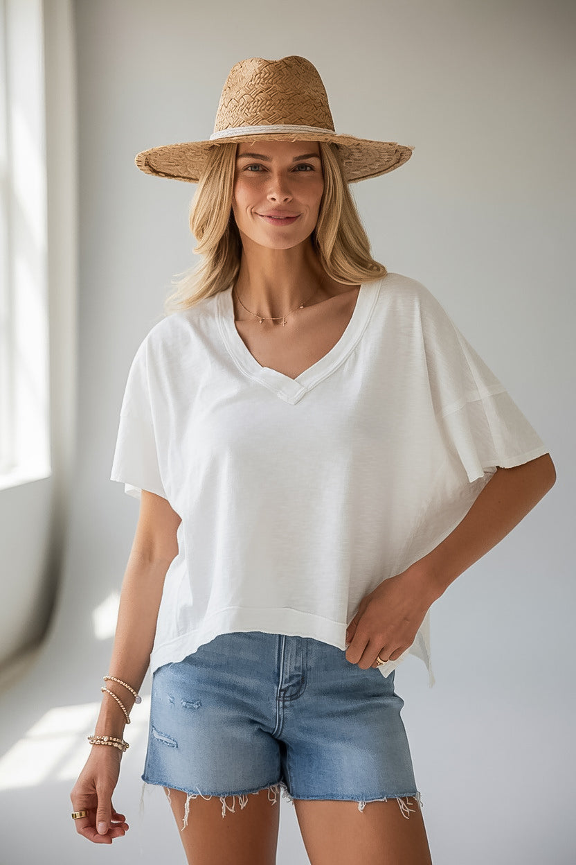 A woman wearing a white short-sleeve V-neck top, light blue denim shorts, and a straw hat, standing in a brightly lit indoor setting.