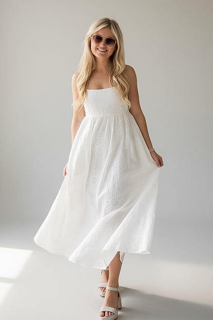 A woman modeling a white strapless midi dress with a gathered waist and flowing skirt, paired with white heeled sandals and sunglasses, against a light gray studio background.