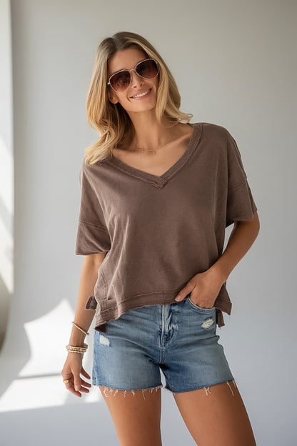 A woman wearing a brown short-sleeve V-neck t-shirt and blue denim shorts, posing in front of a neutral light gray wall.