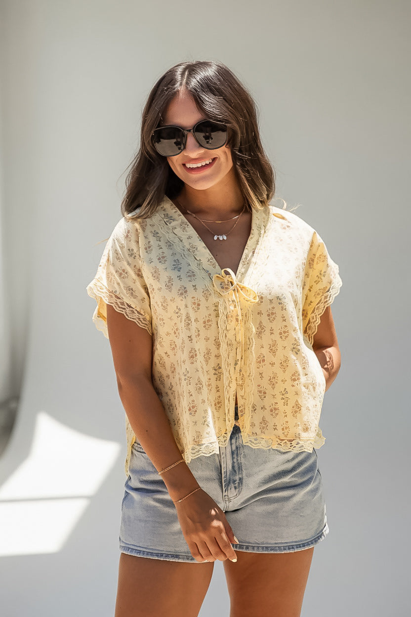 A woman wearing a light yellow floral short-sleeve tie-front blouse with lace trim and denim shorts, standing against a plain light gray wall.
