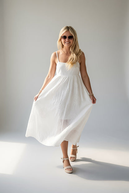 A woman wearing a white sleeveless midi dress and beige strappy heels, standing in a studio with a light gray background.