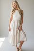 A woman modeling a sleeveless cream-colored dress with a textured knit bodice and a tiered skirt with subtle horizontal stripes, set against a plain light gray studio background.