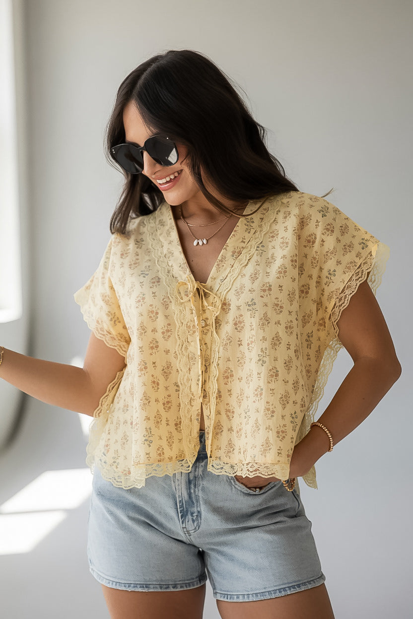 A woman wearing a light yellow short-sleeve lace-trim blouse with a small floral print, styled with denim shorts and sunglasses.
