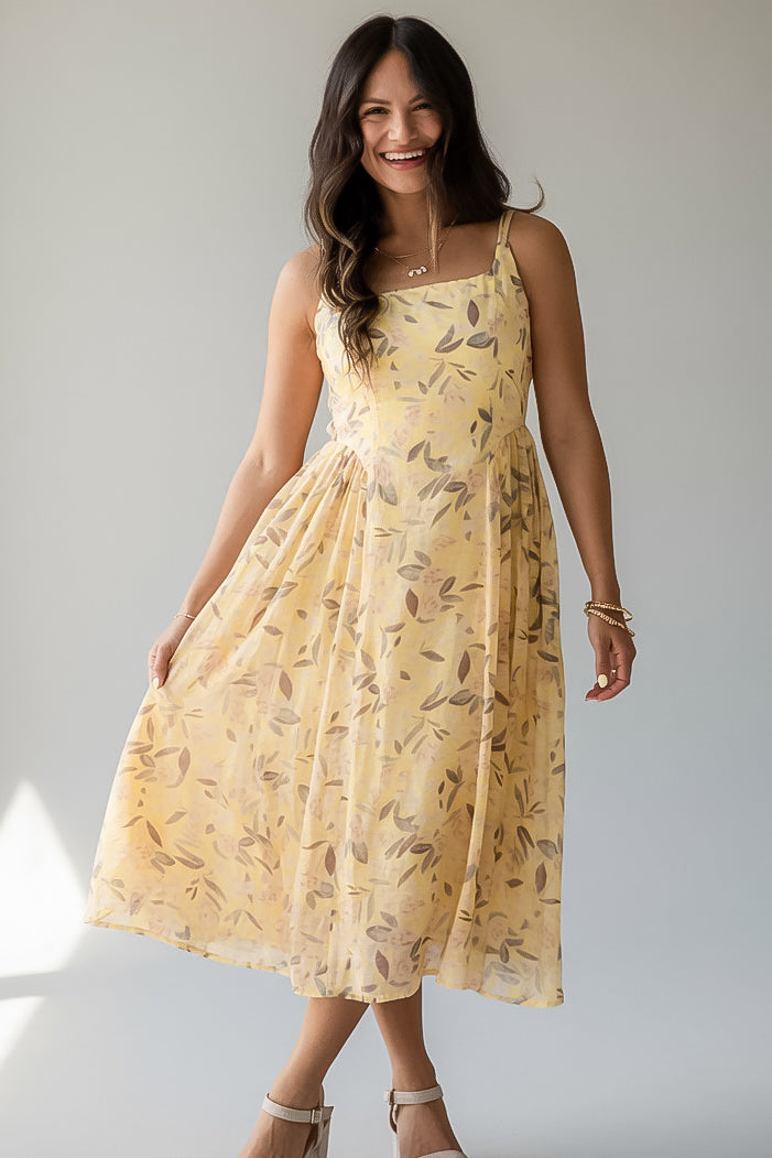 A woman modeling a light yellow sleeveless midi dress with a brown leaf pattern, standing against a neutral light gray background.