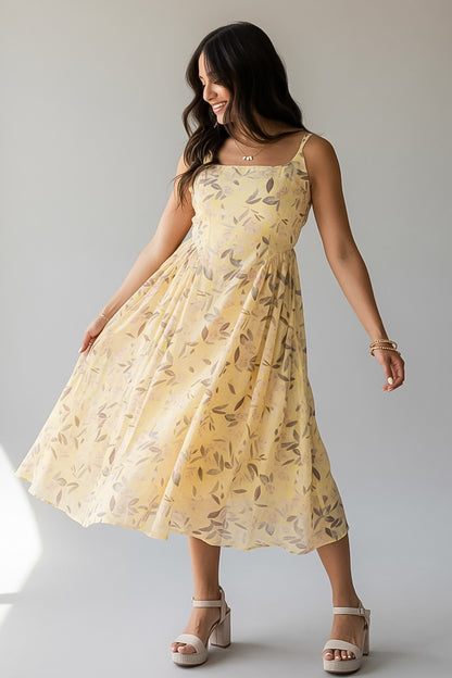 A woman modeling a light yellow sleeveless midi dress with a subtle leaf pattern, shown against a plain light gray studio background.