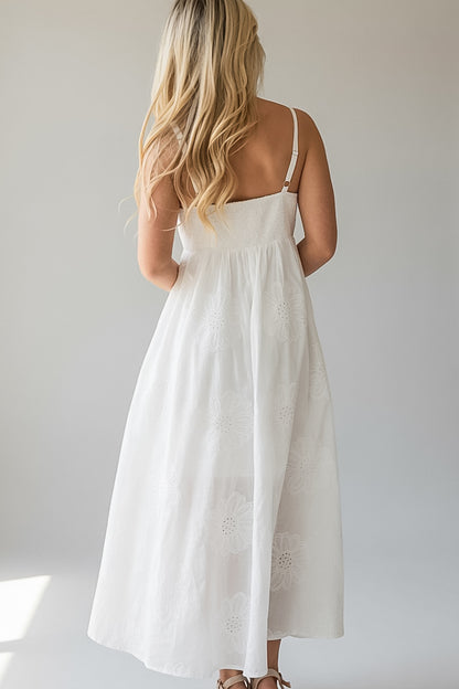 A woman viewed from behind wearing a white midi dress with thin straps and subtle floral embroidery, standing against a plain light gray background.