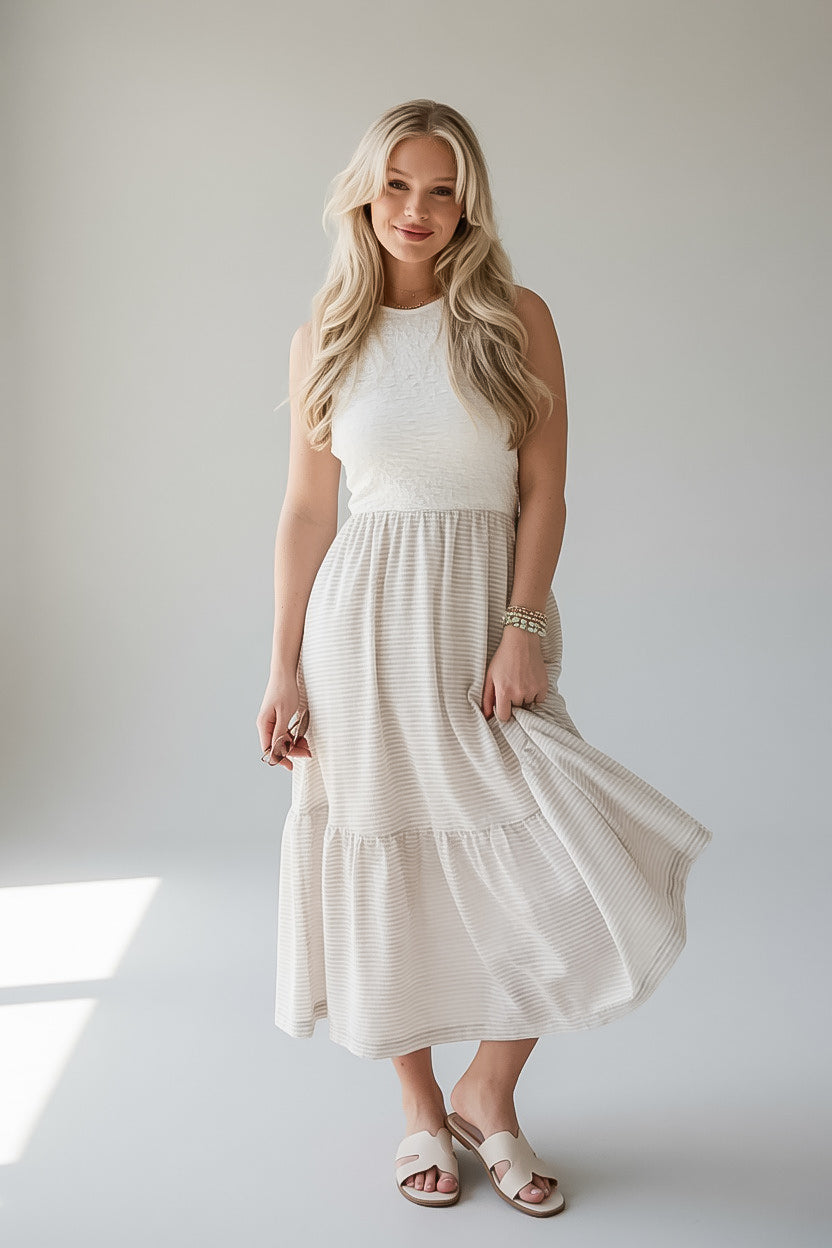A woman modeling a sleeveless cream-colored tiered midi dress with a textured bodice and flowing skirt, paired with neutral slide sandals, set against a light gray studio background.