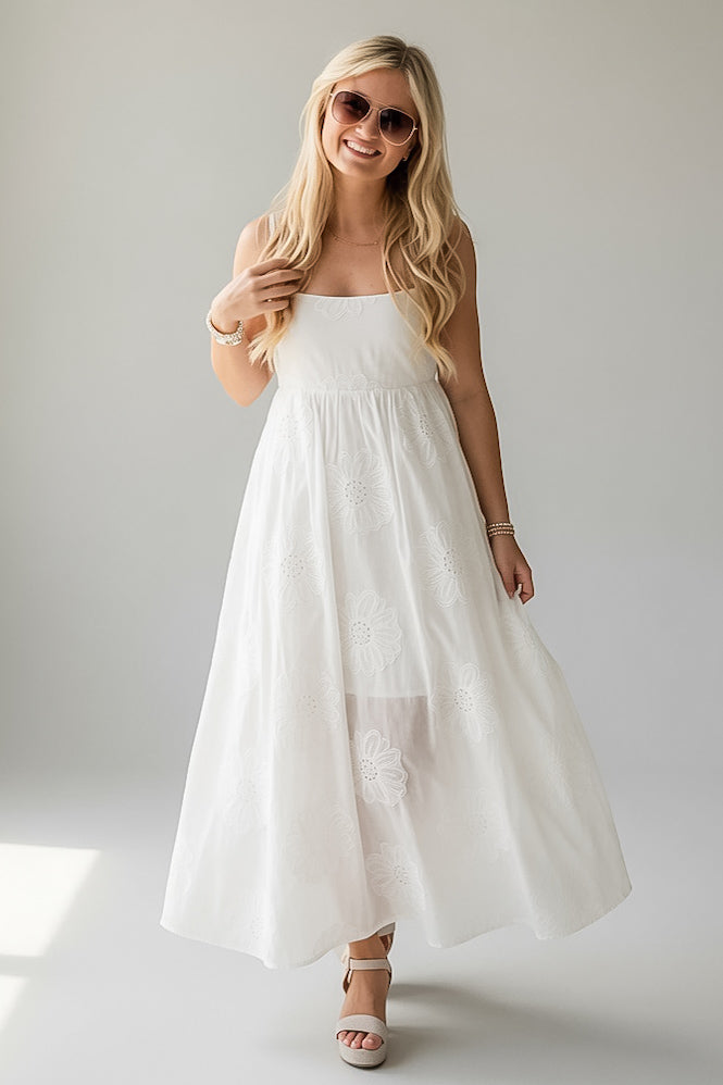 A woman modeling a white sleeveless maxi dress with a gathered bodice and flowing skirt, styled with sunglasses and white heeled sandals against a light gray studio background.