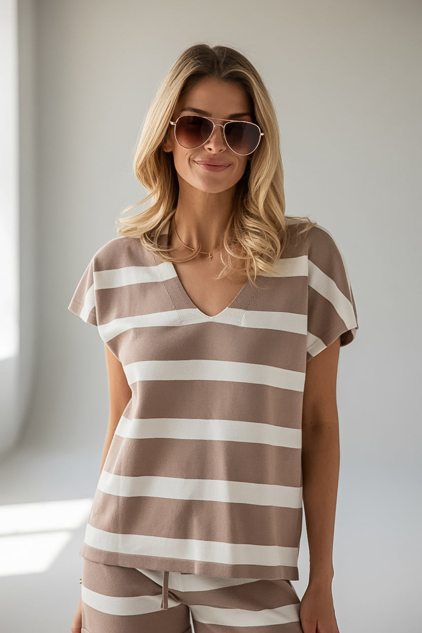 A woman wearing a brown and white horizontally striped short-sleeve V-neck top and matching striped pants, styled with sunglasses, in a brightly lit indoor setting.