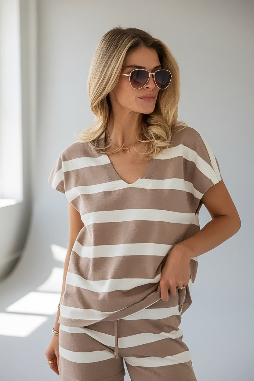 A woman wearing a brown and white horizontally striped short-sleeve V-neck top and matching striped shorts, styled with sunglasses, in a brightly lit room.