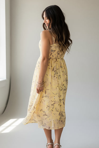 A woman wearing a light yellow sleeveless midi dress with a brown leaf pattern, photographed from behind in a bright indoor setting.
