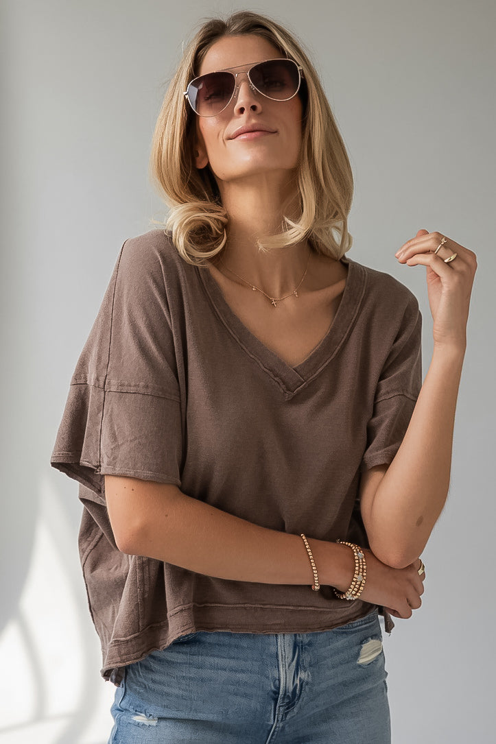 A woman wearing a brown short-sleeve V-neck t-shirt and blue jeans, styled with sunglasses, bracelets, and a necklace, posing against a neutral gray background.