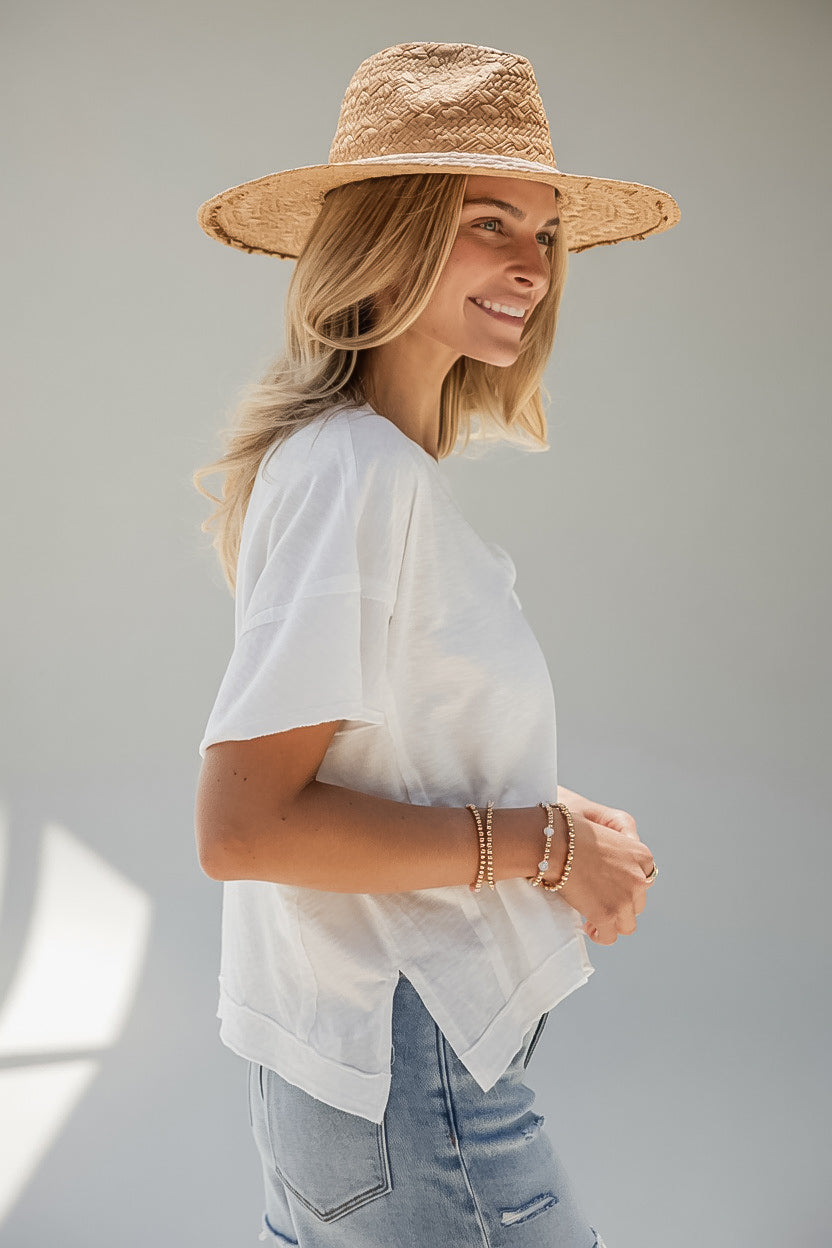 A woman wearing a straw hat, a loose white short-sleeve top, and light blue jeans, shown in profile against a plain light gray background.
