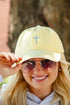 A close-up lifestyle shot of a smiling blonde woman wearing a pale yellow baseball cap with a blue embroidered cross on the front, gold-framed sunglasses, and a gray hoodie, with a blurred tree trunk in the background.